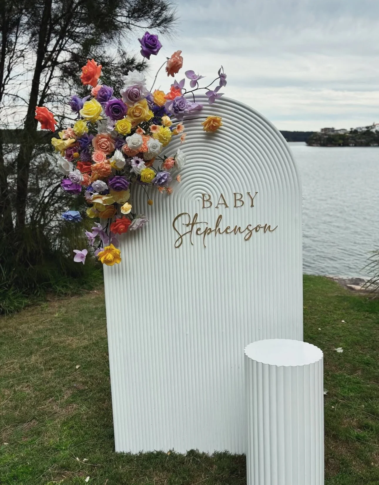 White arched sign with gold lettering saying 'Baby Stephenson' decorated with a large bouquet of colorful flowers, situated on a grassy area near a body of water with trees and buildings in the background.
