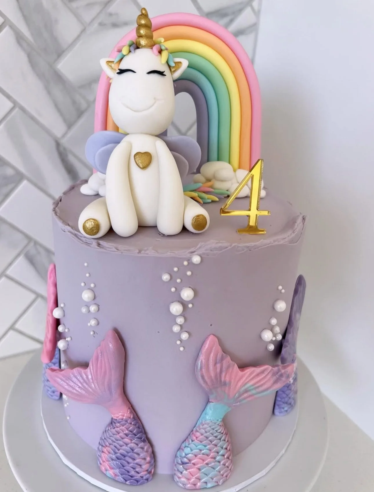 A pastel-colored unicorn-themed birthday cake with a unicorn figurine, rainbow, and mermaid tails, decorated with pearls and a gold number 4 candle.