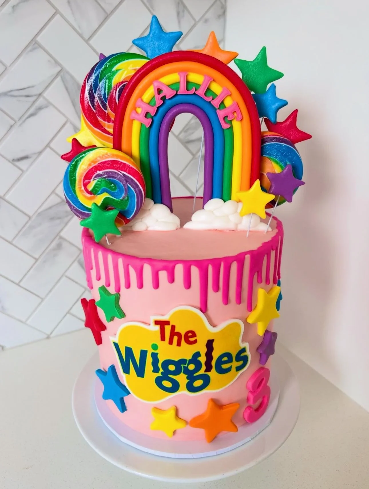 Pink birthday cake with rainbow drip decoration, colorful star and swirl lollipop toppers, and a rainbow-shaped topper with the name "Halie" on top. The front of the cake has a yellow sign that says "The Wiggles" and multicolored stars around its sides.