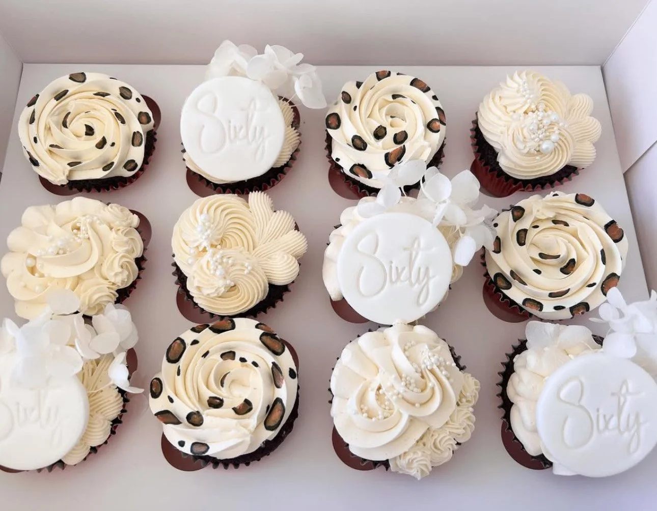 A box of twelve cupcakes decorated with white and dark chocolate frosting, some with leopard print designs, and some with white edible toppers reading 'Sixth' and 'July', adorned with white edible pearls and decorative white flowers.