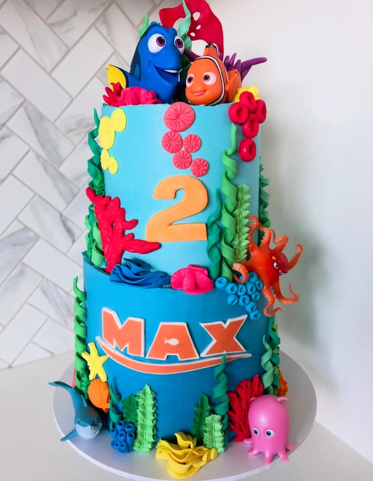 A three-tiered birthday cake with an underwater ocean theme, decorated with colorful fondant sea creatures including Nemo, Dory, an octopus, a shark, and a pink octopus, with the name 'Max' and the number 2, indicating a second birthday.