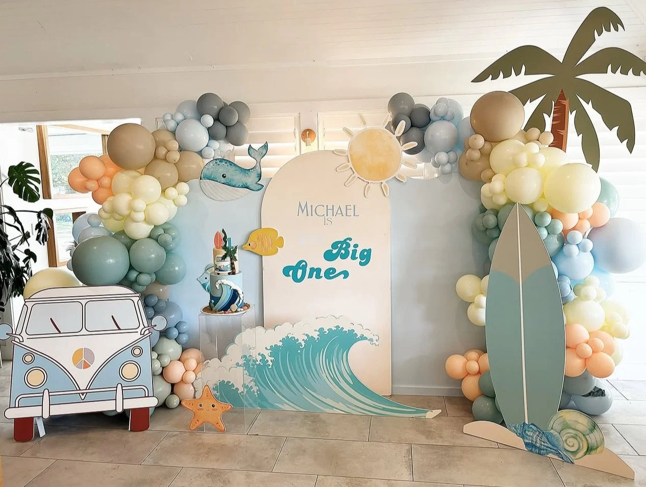 Beach-themed birthday party decoration with a balloon arch featuring pastel colors, a surfboard, wave illustration, a cartoon van, a whale, sun, palm tree, and sea creature cutouts.