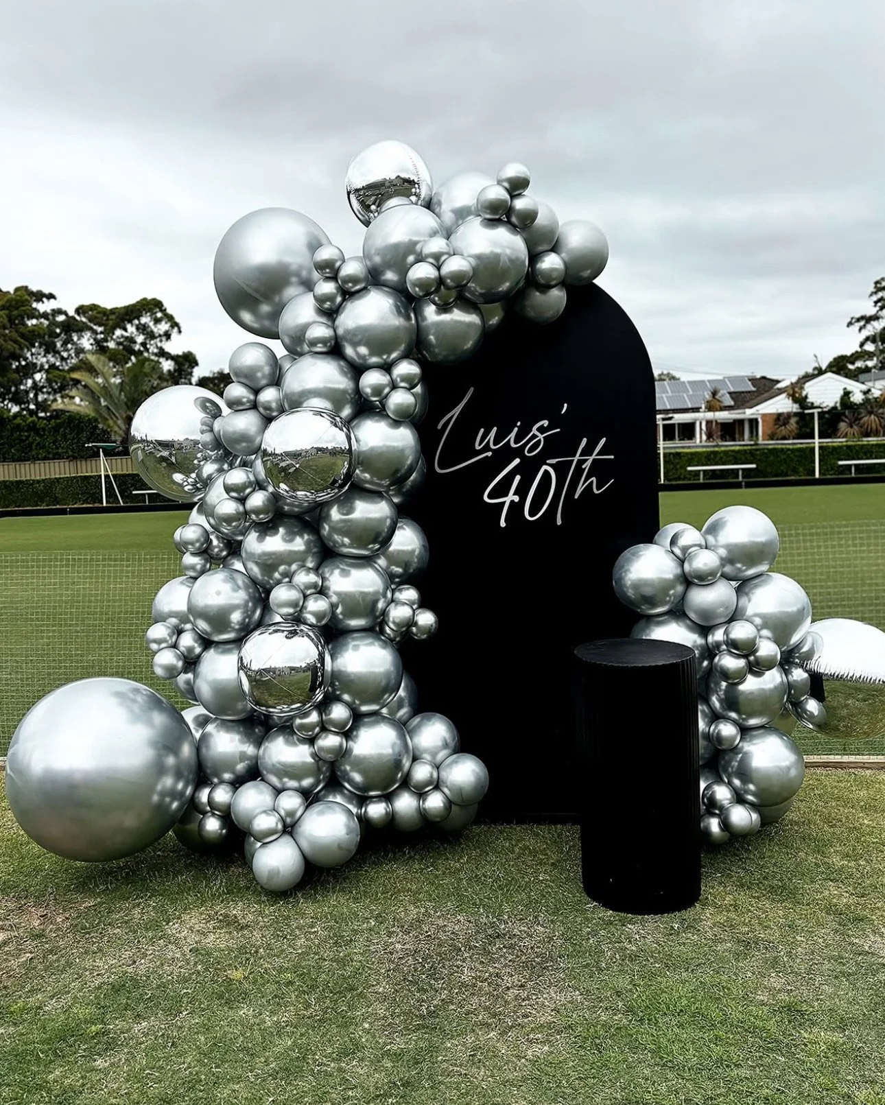 Decorative balloon arrangement surrounding a black board with white cursive text reading "Luis' 40th" on a lawn, with a tennis court and cloudy sky in the background.