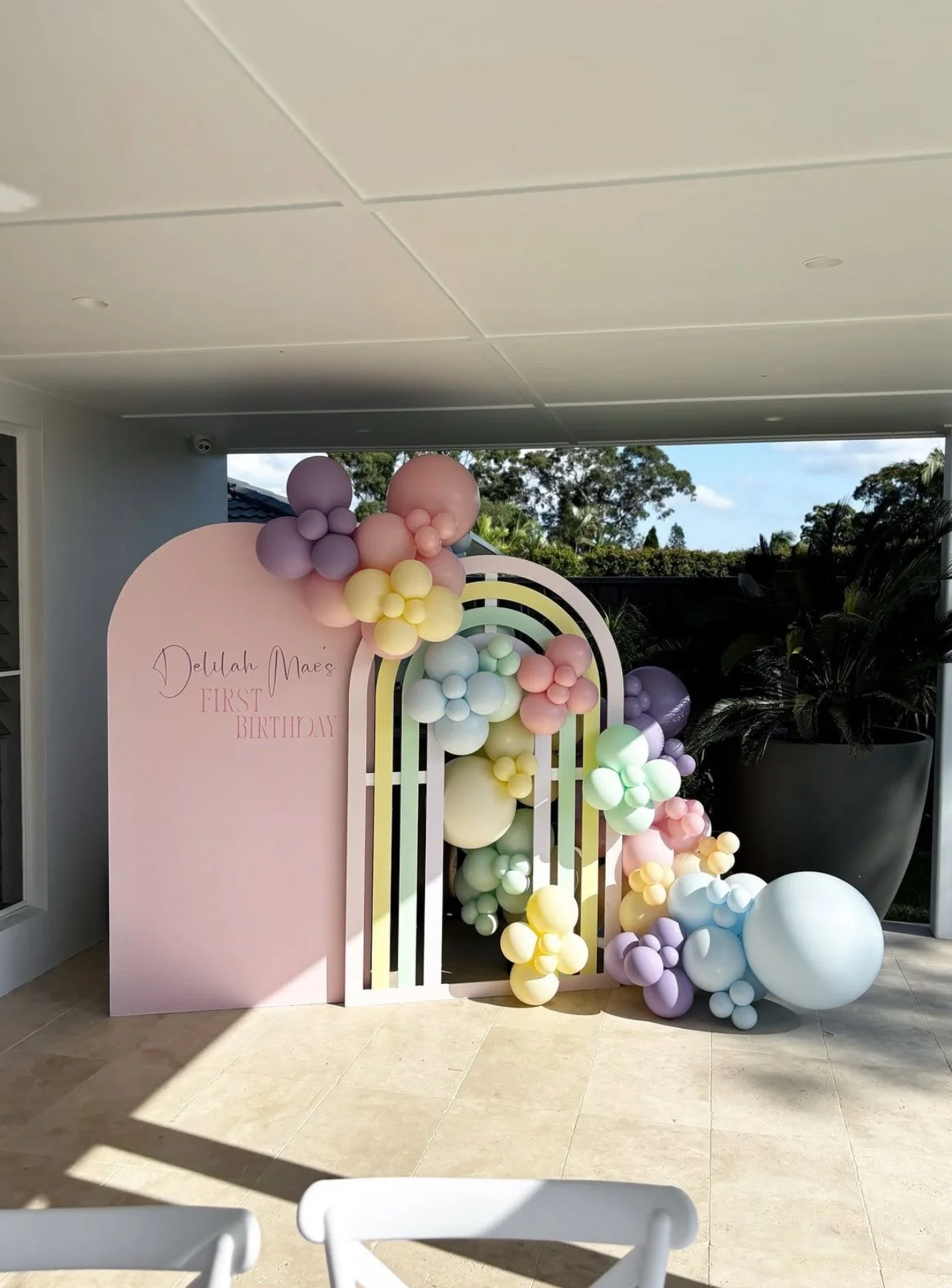 Decorative pastel-colored balloons arranged around a pink and white wooden arch with a sign that reads 'Delilah Mae's First Birthday.'