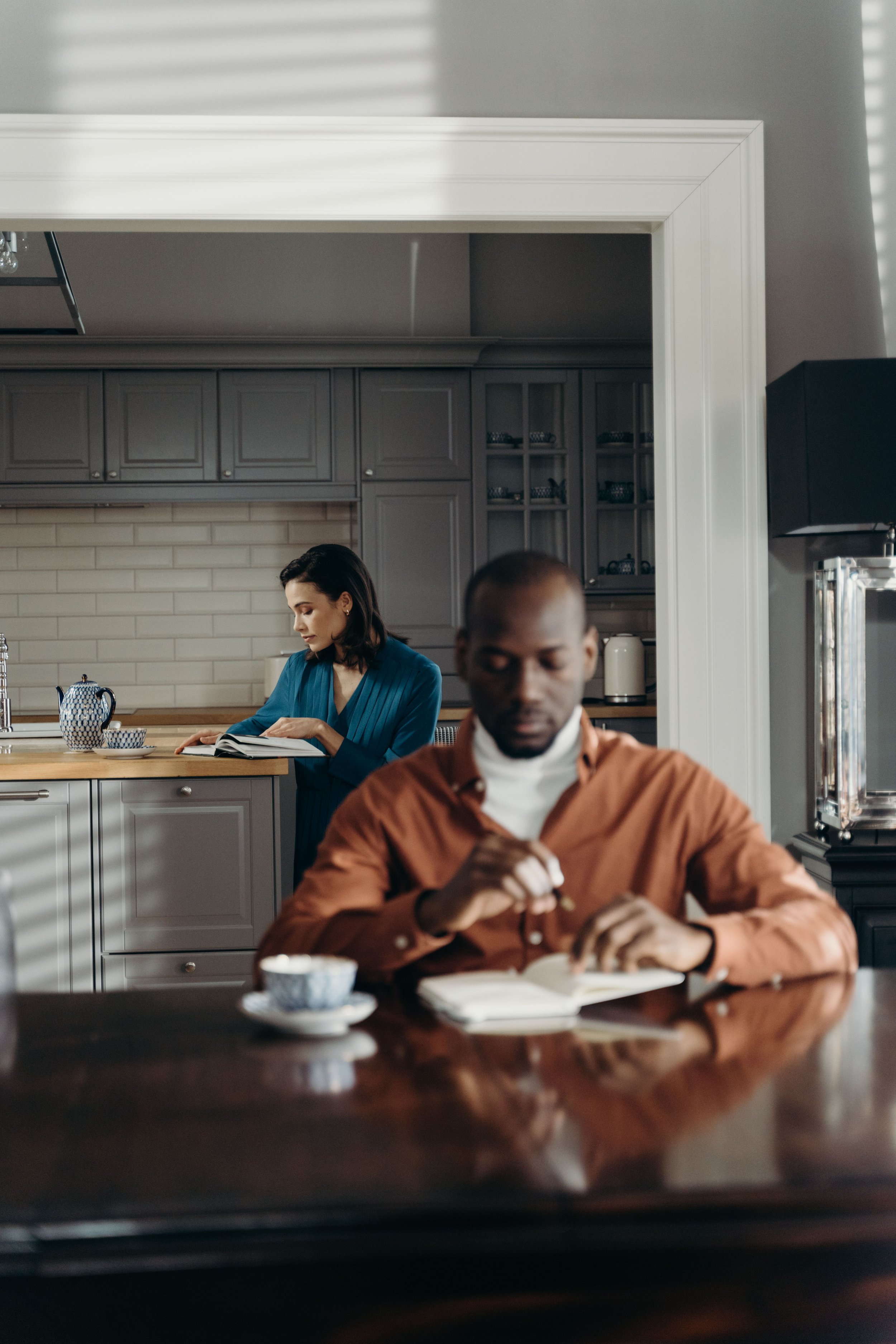 A couple reading separately in the same room, one at the table and one at the bar, representing the parallel lives that many empty nest couples find themselves living when children leave home