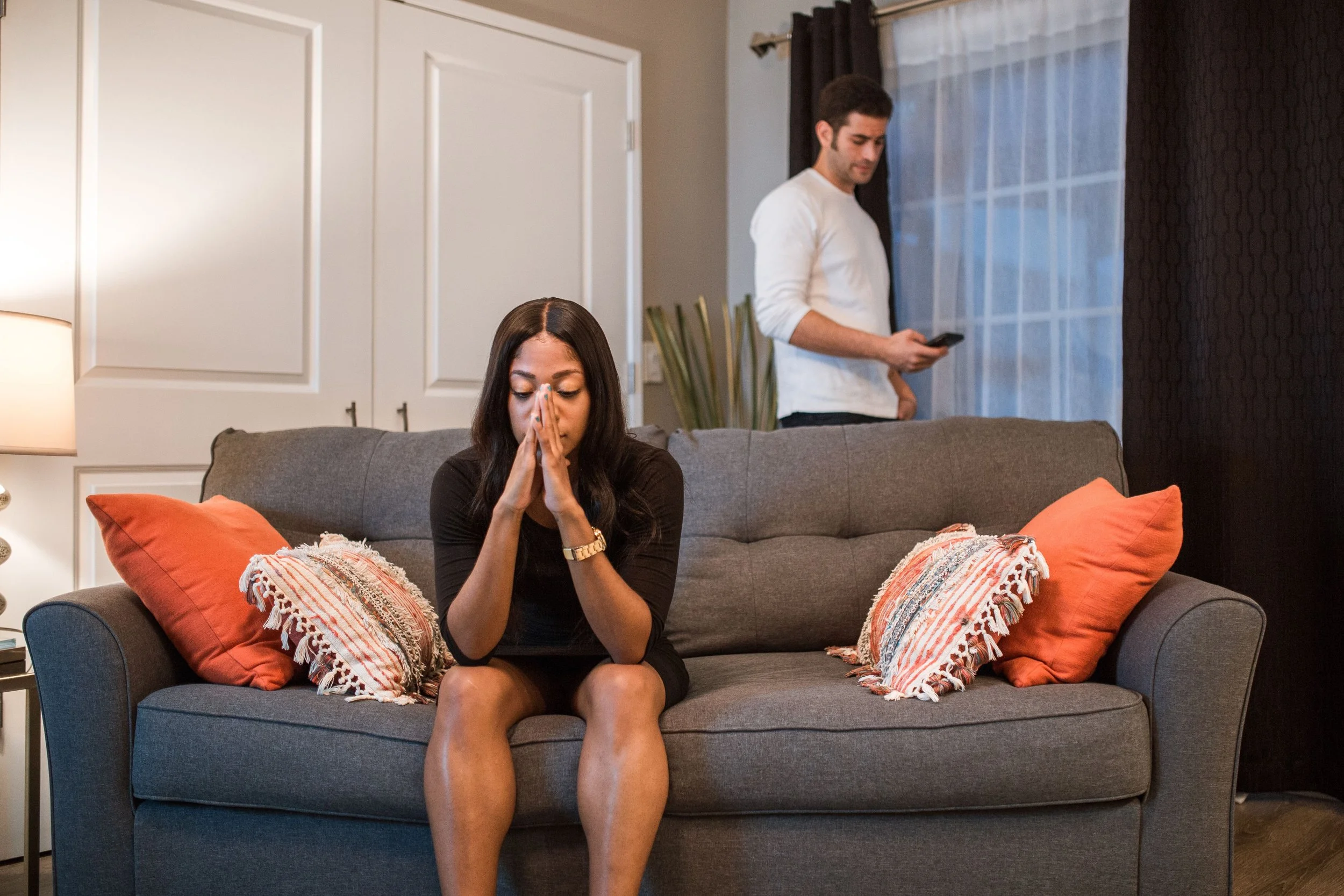 Couple in a living room with one person using a phone behind the couch while the other sits looking down.