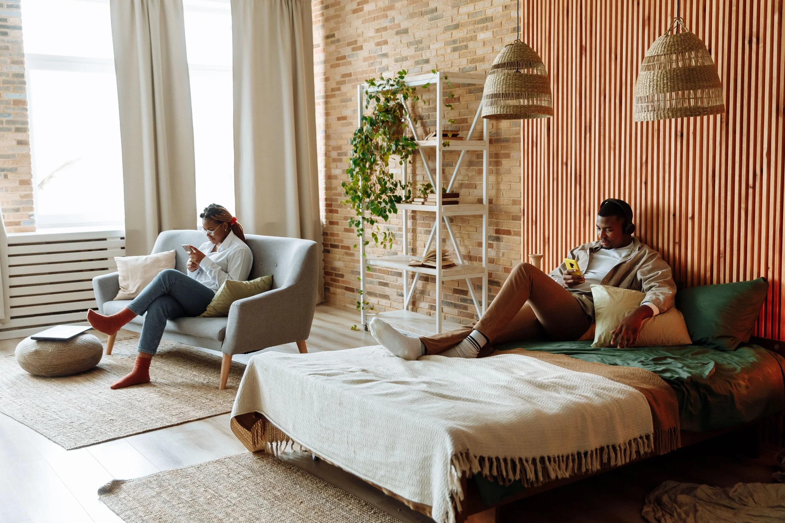 A couple sits apart in a warmly lit room — man on the bed, woman on the couch, each on their phone — representing the disconnection that trauma-informed couples therapy in Austin, TX can help address.