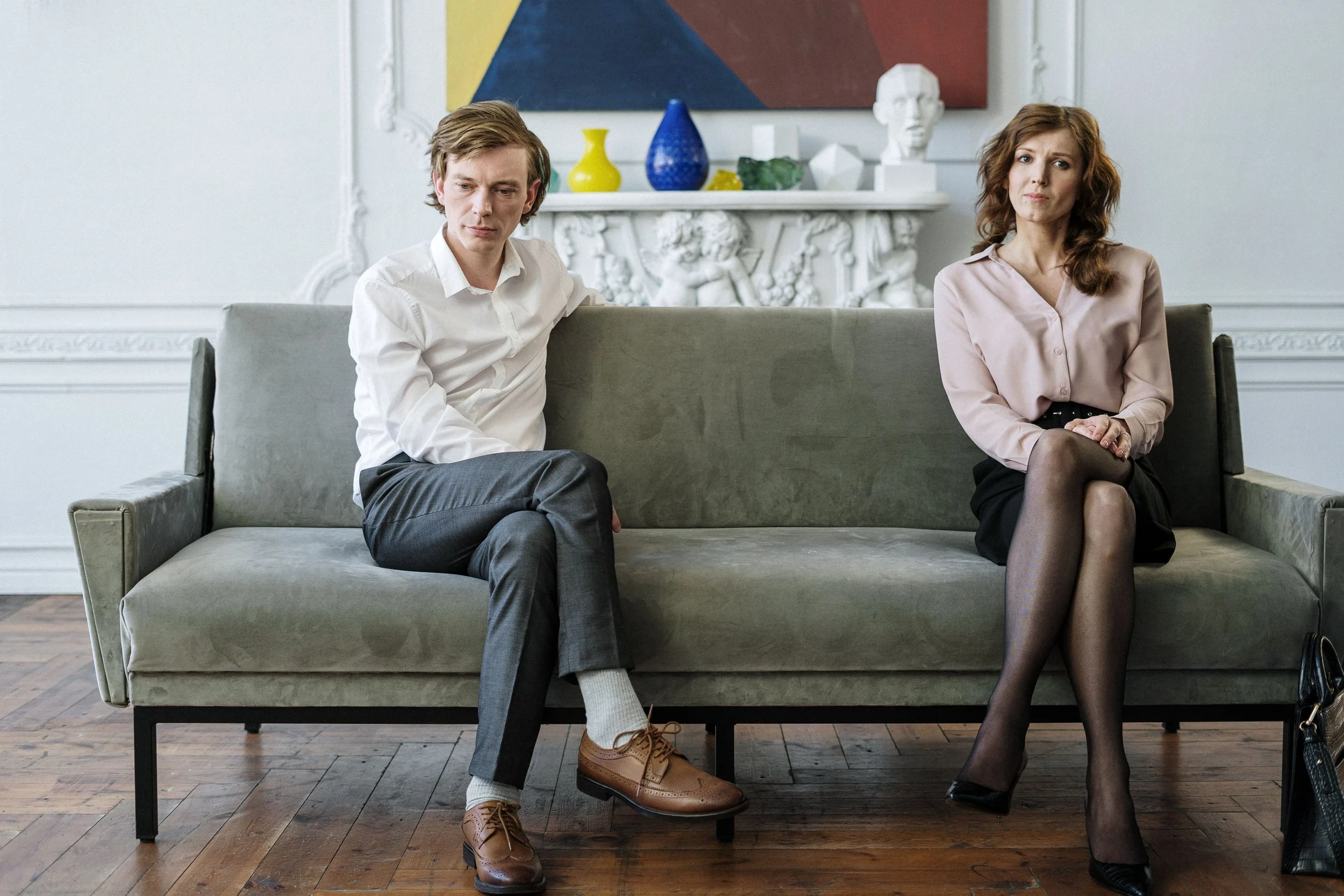 Couple sitting apart on a couch, both looking away, reflecting the emotional distance and disconnection that characterizes the early stages of affair recovery therapy