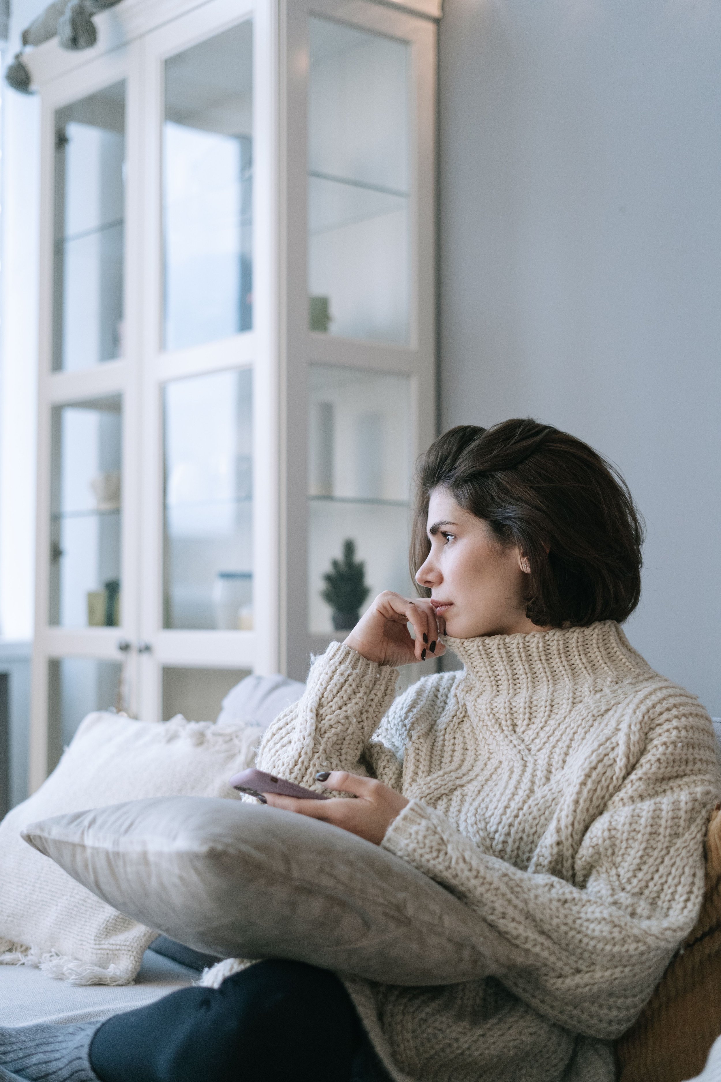 A woman sits on a couch in natural light holding a phone while looking out the window, reflecting a quiet everyday moment of inward attention and reflection.