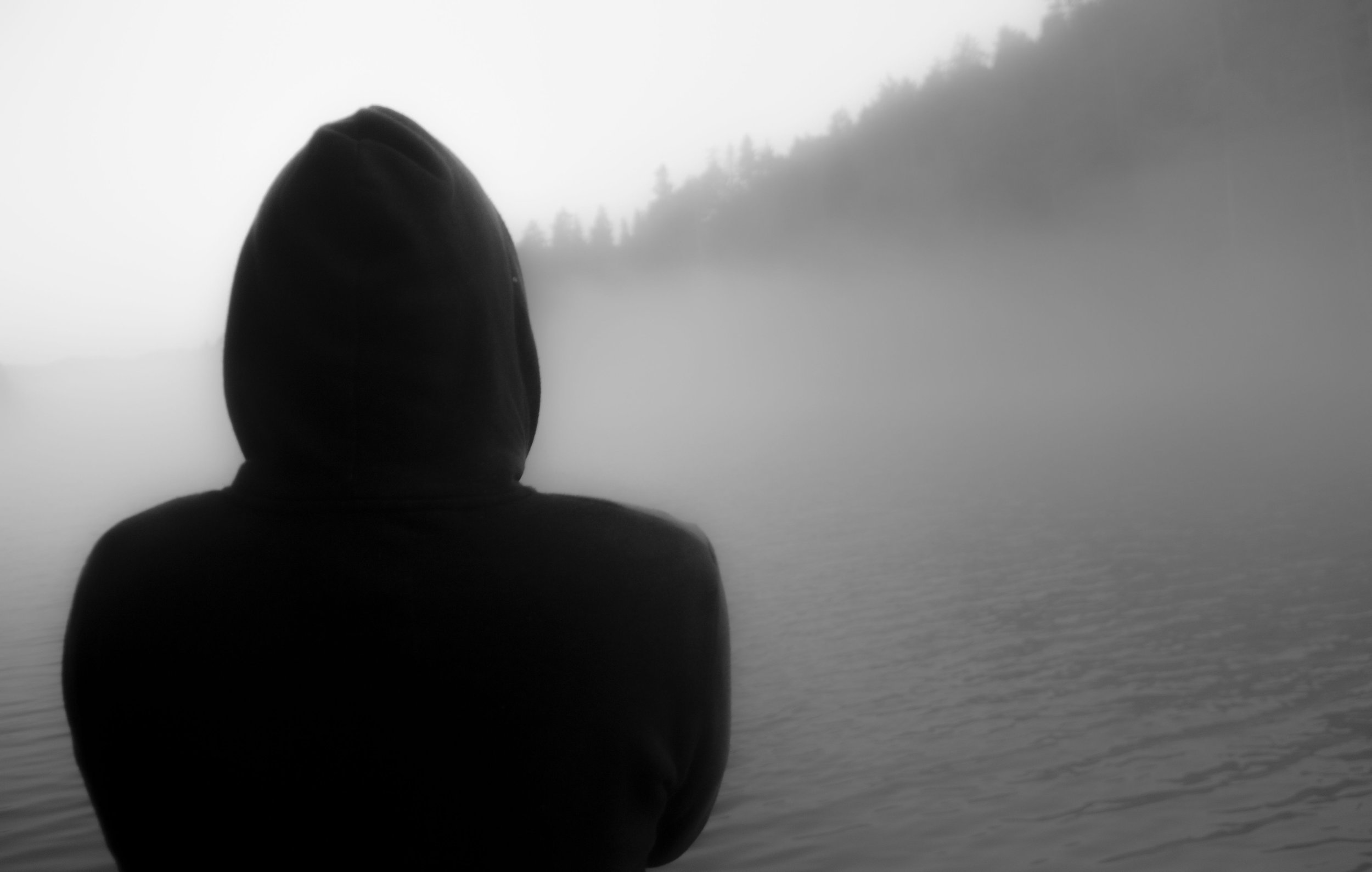 A person in a hoodie standing at the edge of a foggy body of water, representing the disorientation and grief of betrayal trauma and the process of finding solid ground again