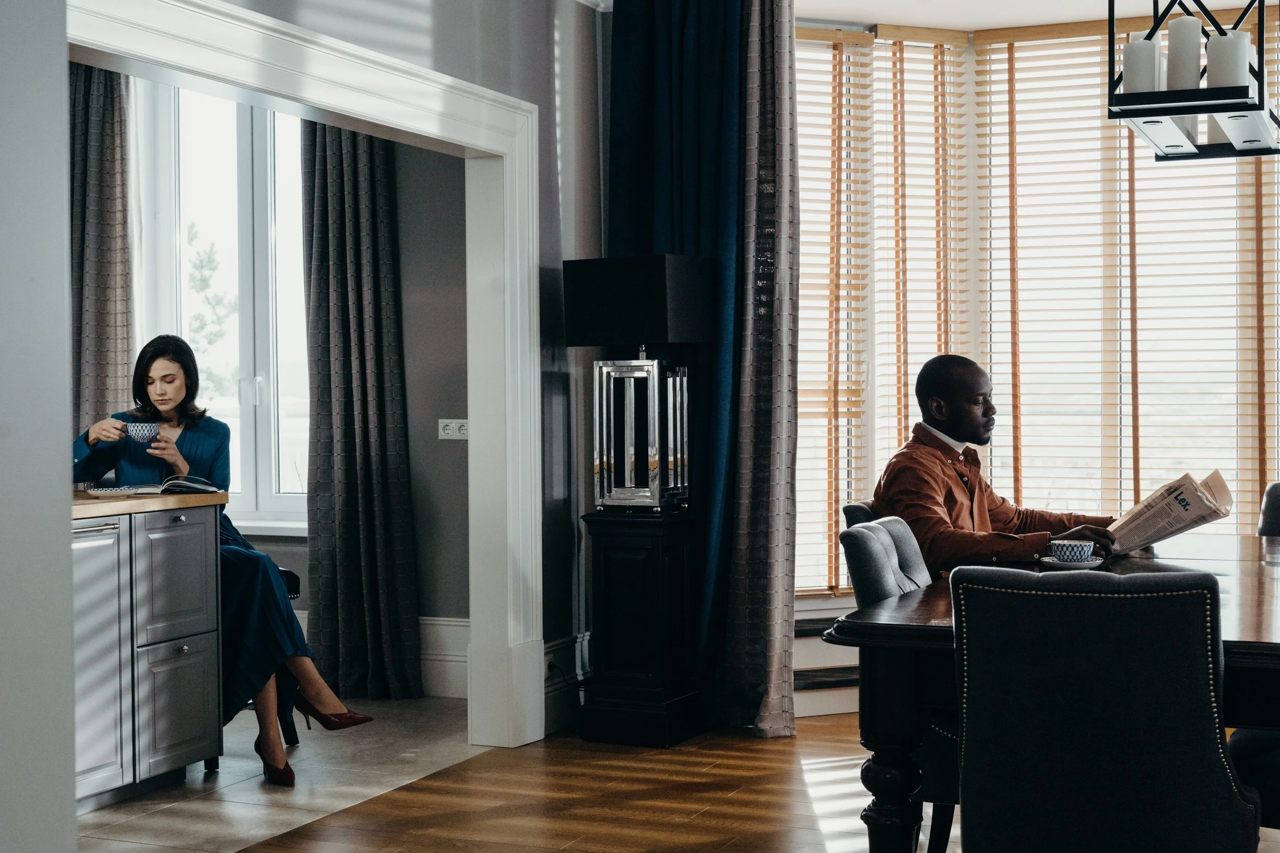 Couple sitting apart in living room, woman drinking coffee reading and man reading newspaper, representing emotional distance in relationships affected by PTSD