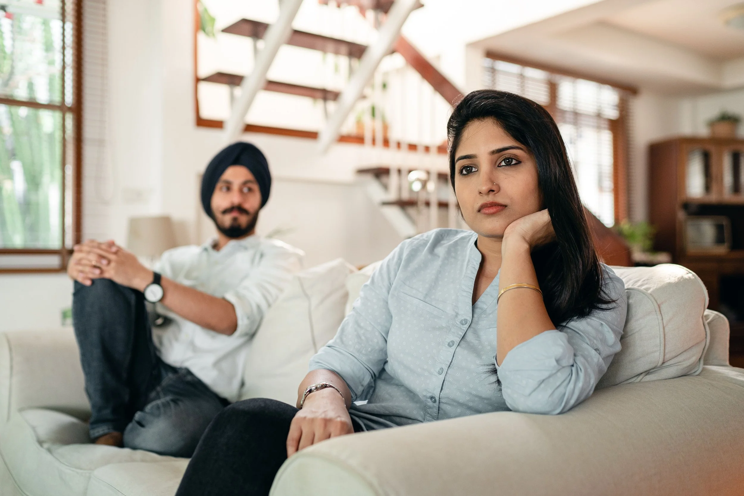 A couple sits at opposite ends of a couch; a woman stares into the distance in the foreground while a man looks toward her from the background, illustrating the emotional distance couples face in affair recovery therapy