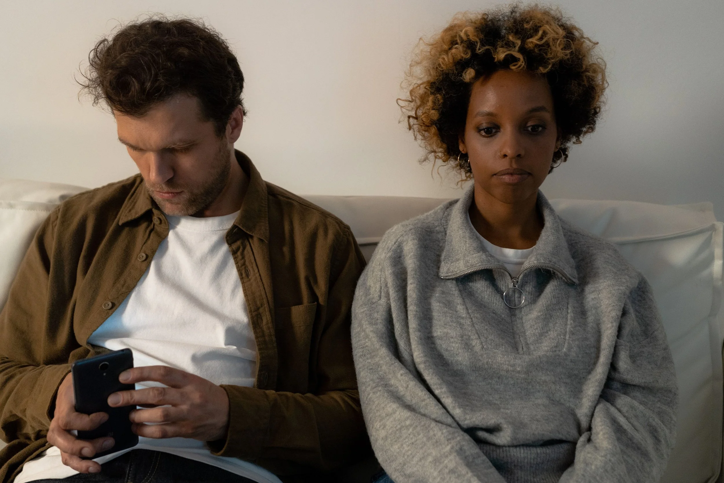 A couple in bed showing emotional distance, as one partner focuses on a smartphone while the other looks away, reflecting digital distraction in intimate relationships.