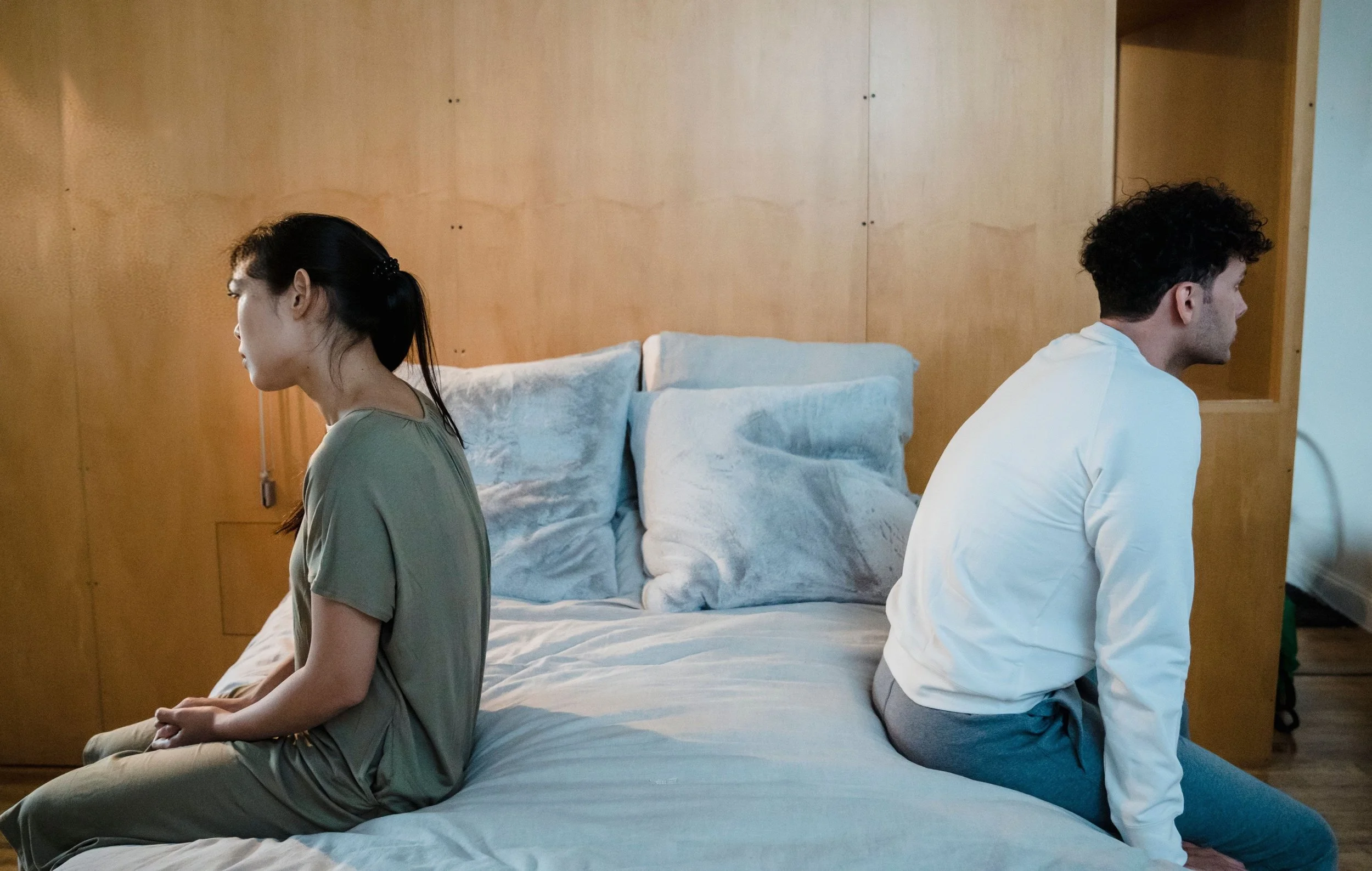 Couple sitting on opposite sides of a bed, emotionally distant, representing the pain of betrayal trauma and the beginning of affair recovery therapy.
