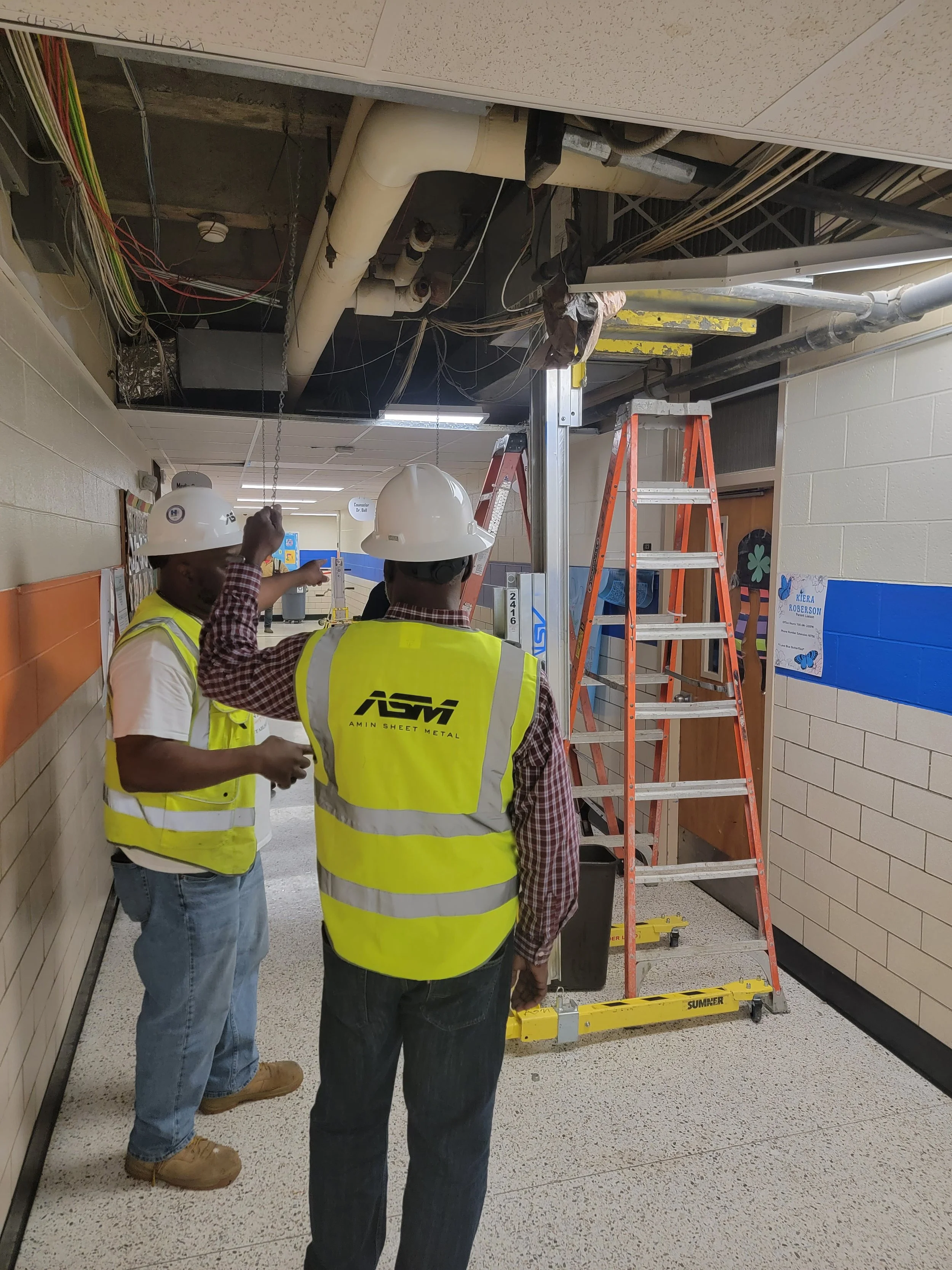 Two workers in safety vests and helmets inspecting ceiling infrastructure in a hallway with ladders and construction tools.