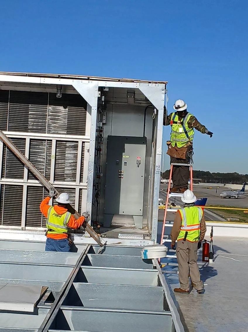 Workers in safety vests and helmets installing or inspecting equipment on an airport tarmac with airplanes in the background.