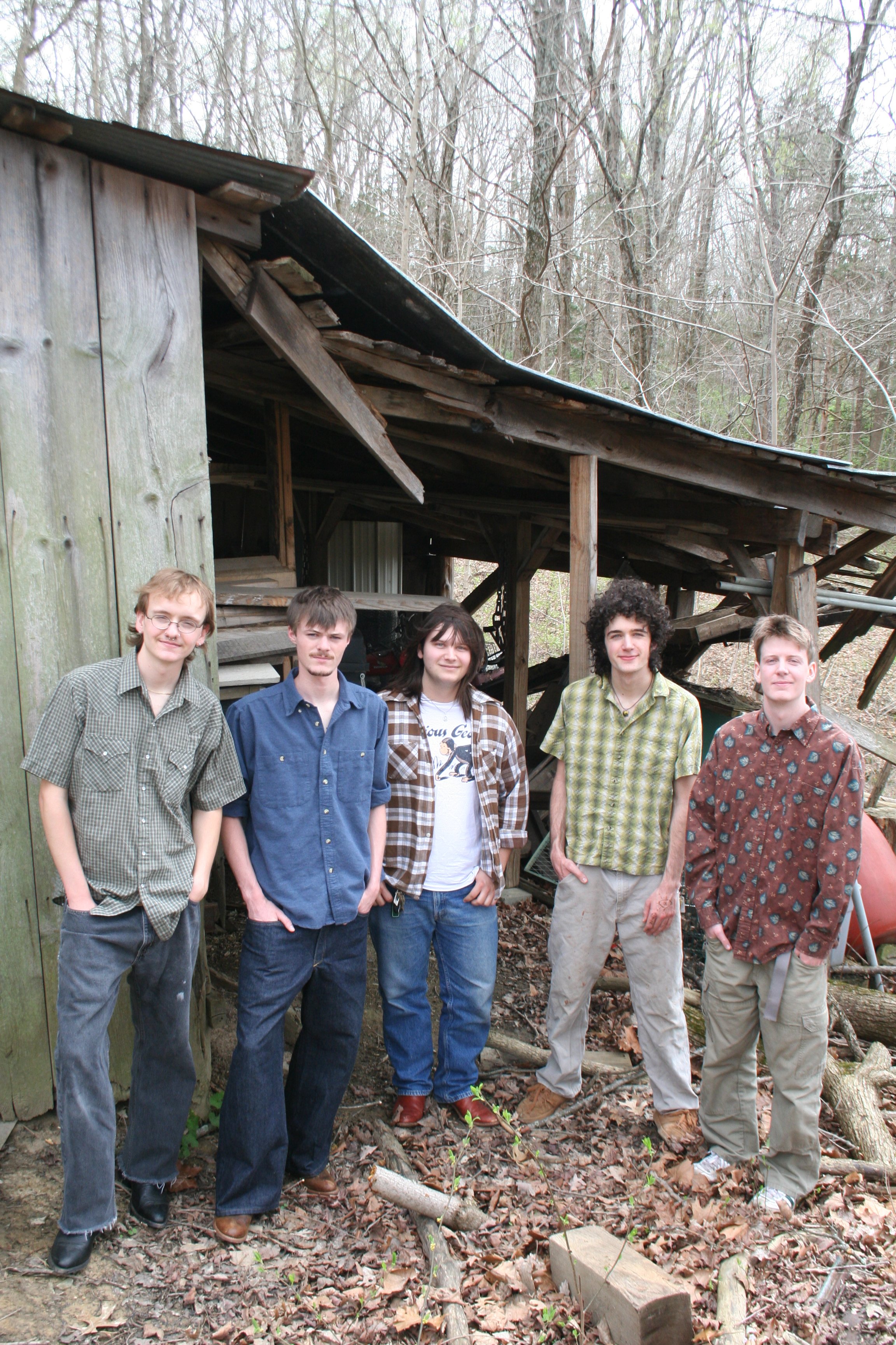 Six young adults standing outdoors near a weathered wooden shed in a wooded area, dressed casually, with trees and fallen leaves in the background.