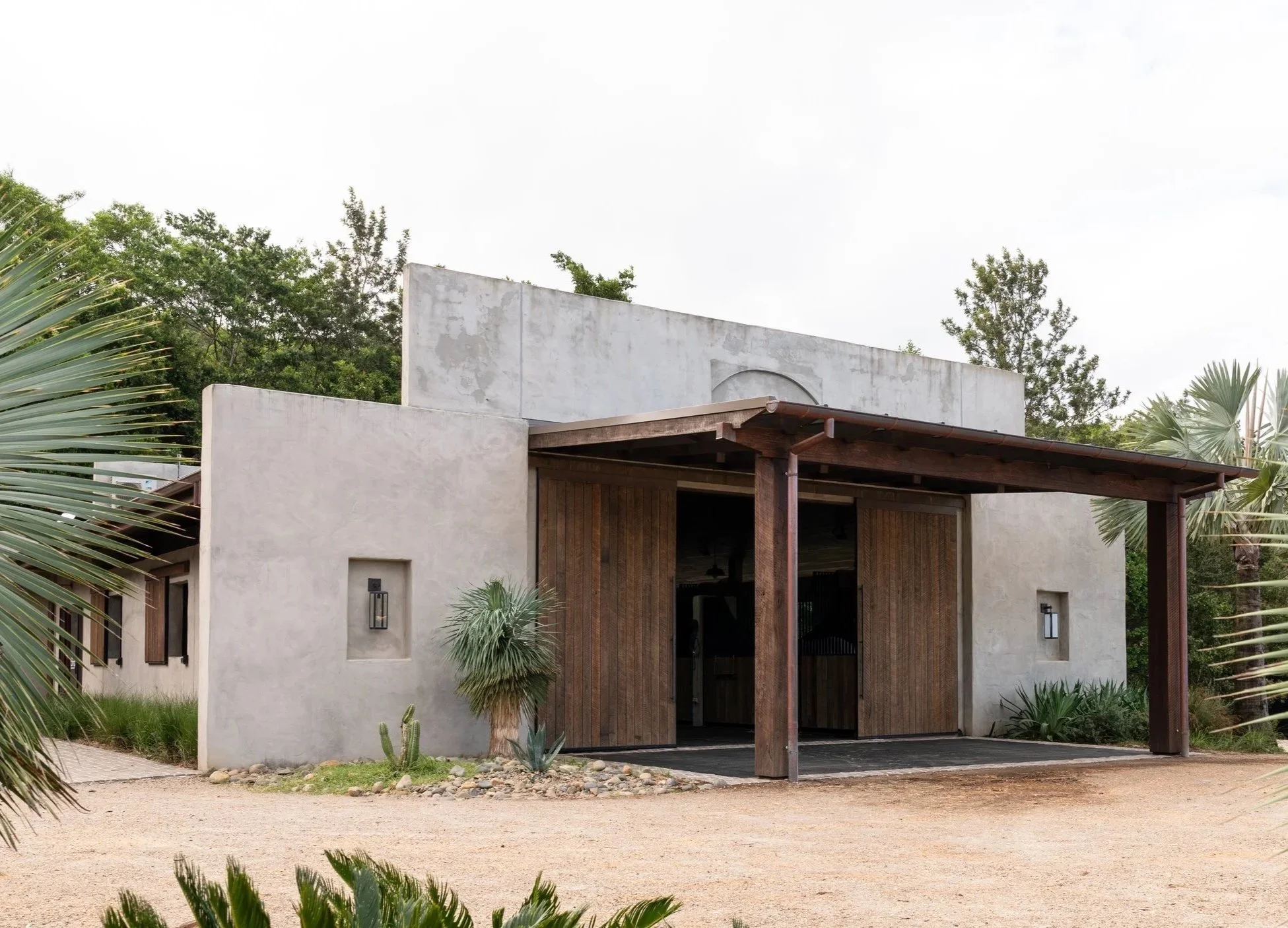 Exterior view of a modern stucco building with wooden accents surrounded by desert landscaping, including palm trees and cacti.