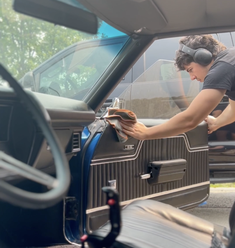 A young man wearing headphones is cleaning the dashboard of a vintage car with a cloth.