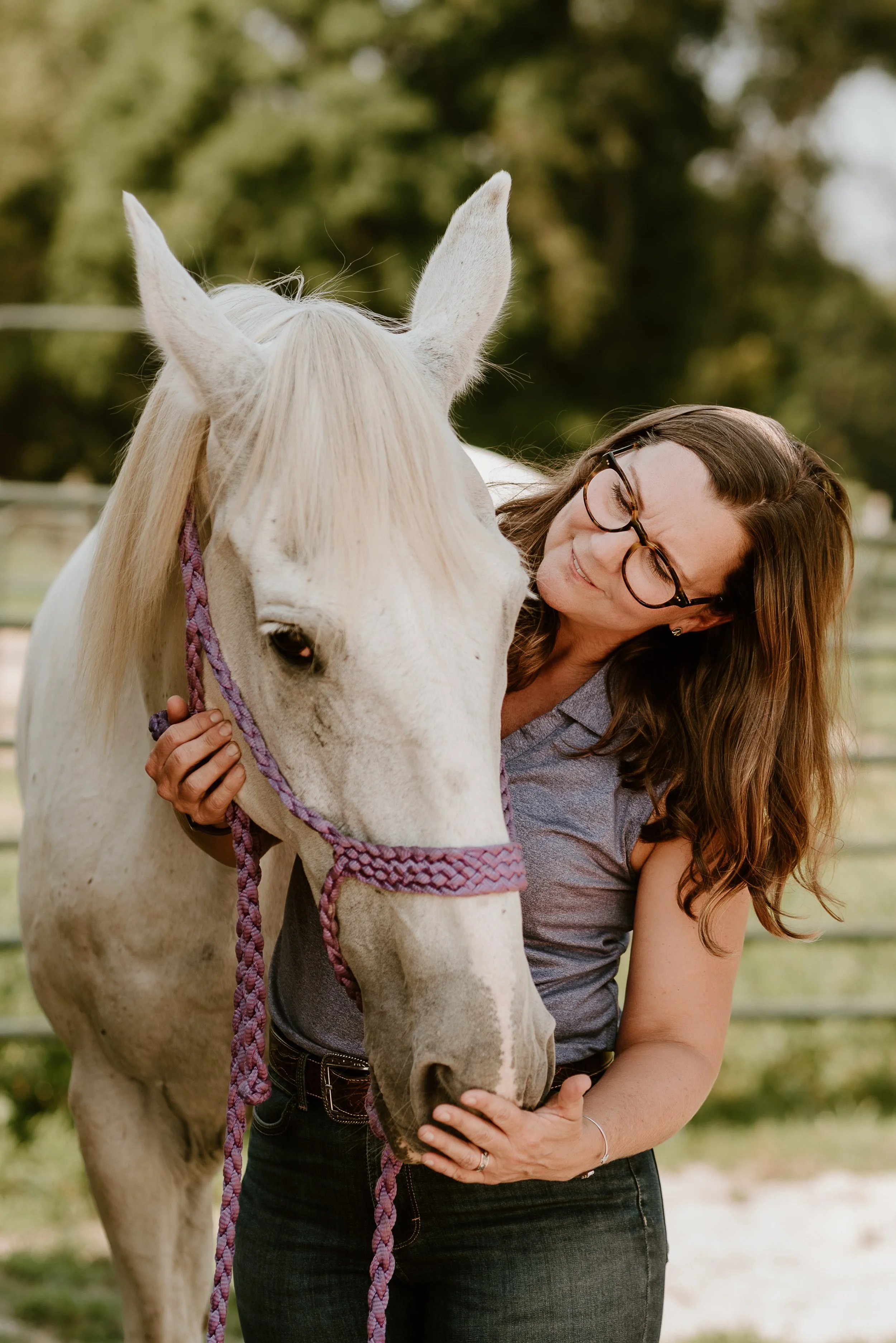 April Schaeffer hugging a white horse wearing a purple halter, outdoors in a green fenced area.