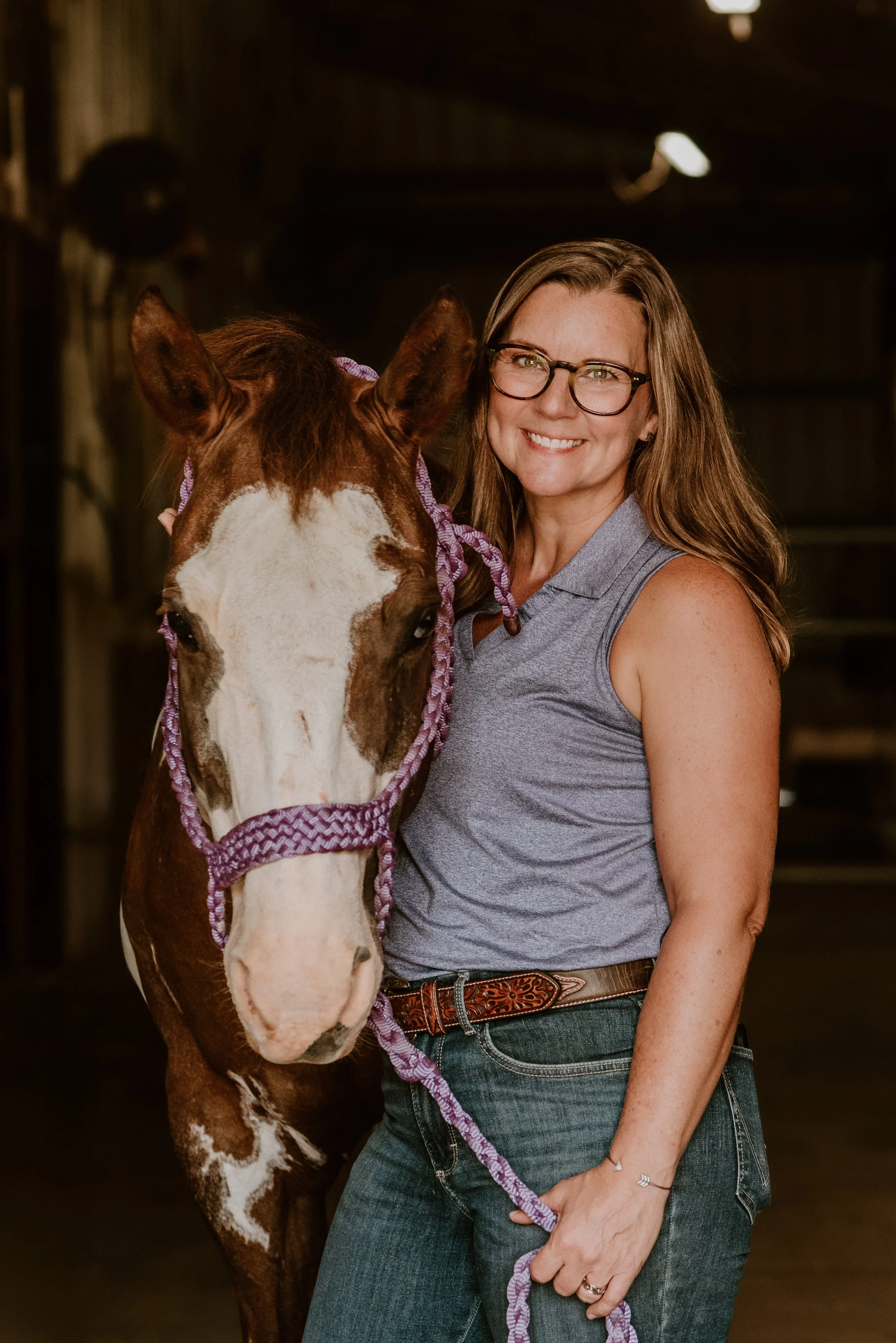 April Schaeffer smiling with glasses beside a brown and white horse in a barn.