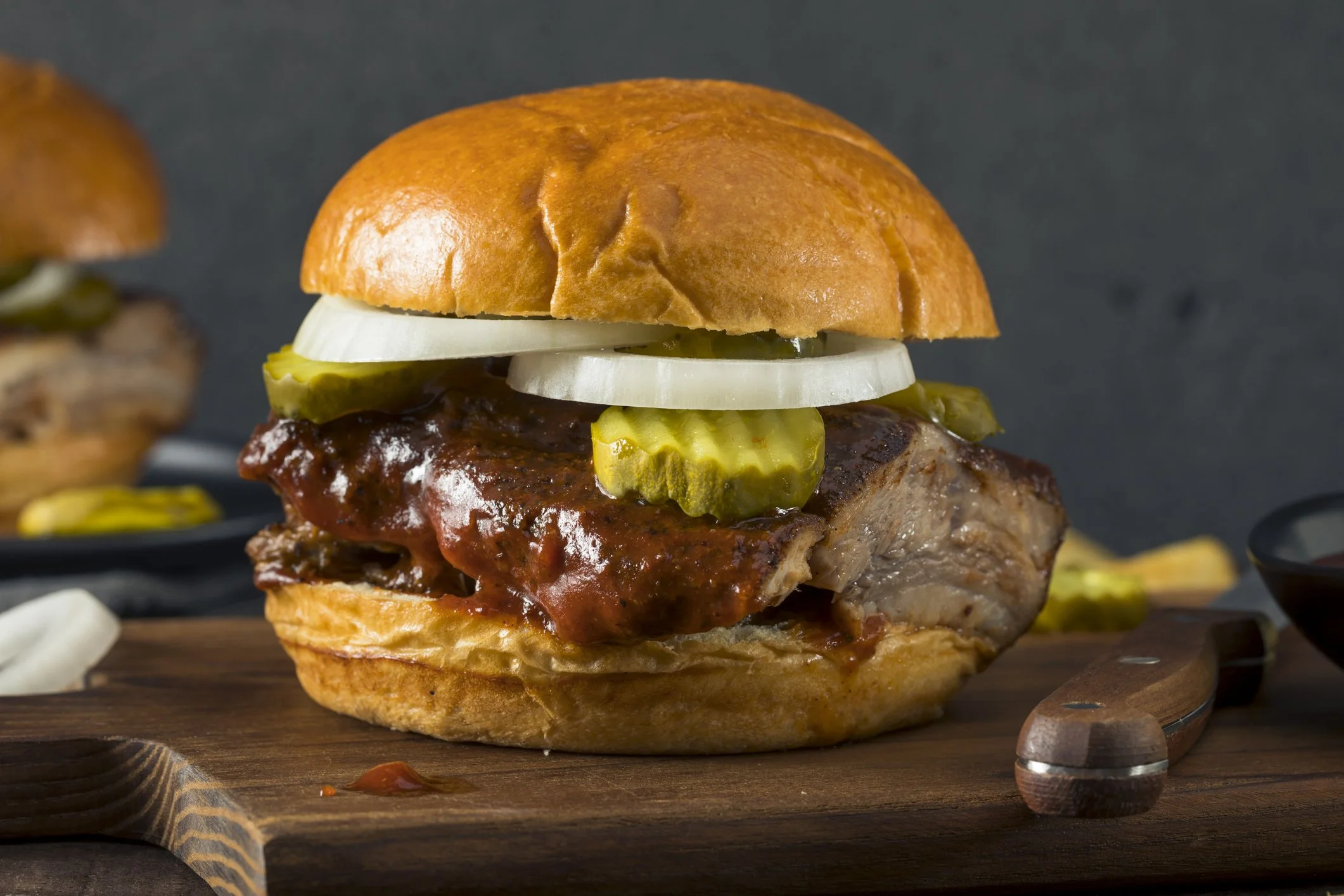 Close-up of a juicy burger with pickles, onion, and barbecue sauce on a brioche bun, served on a wooden board.