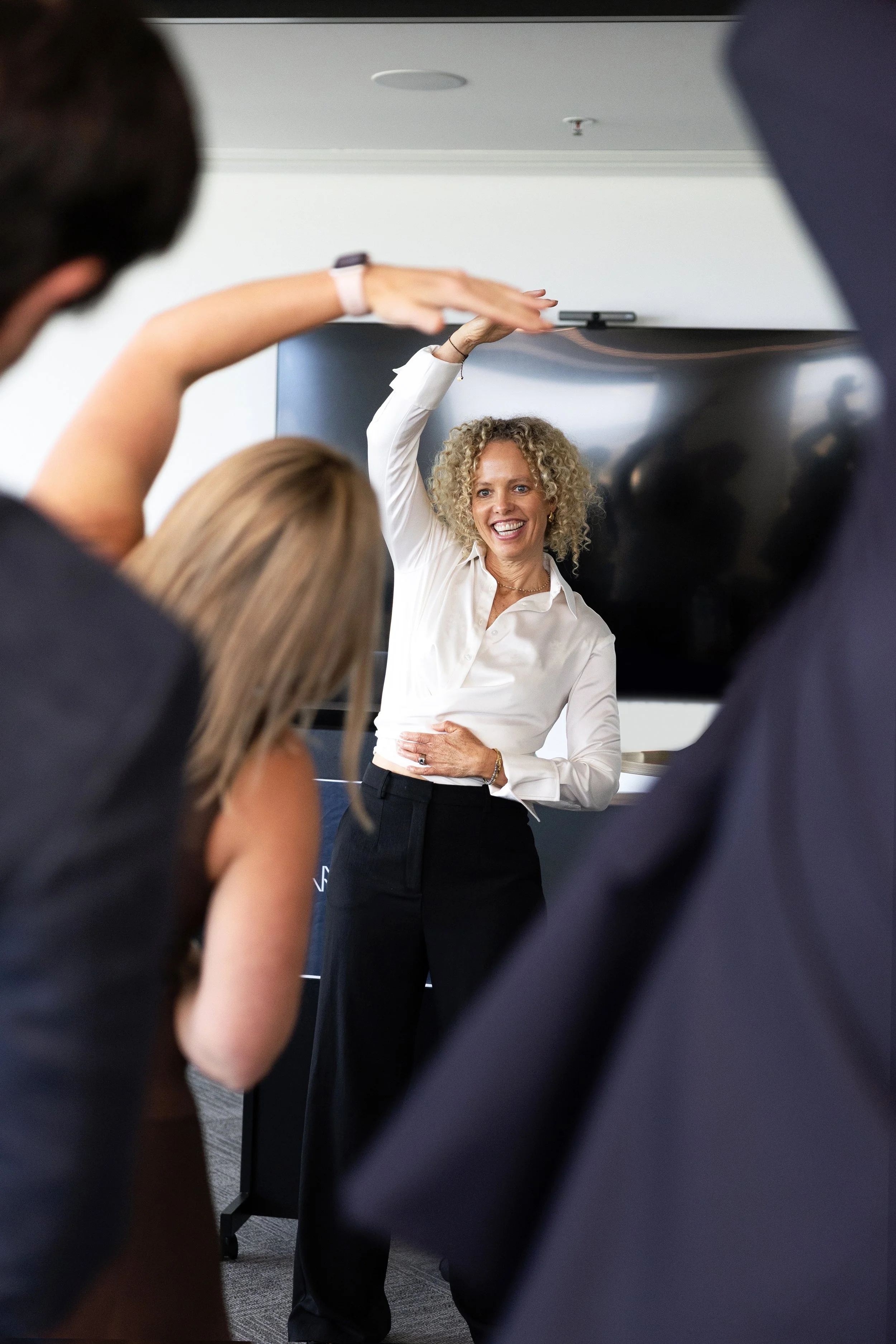 A woman with curly blonde hair wearing a white blouse and black pants is smiling and raising her right hand. She is in a room with a large screen behind her, surrounded by other people, some partially visible and blurred in the foreground.