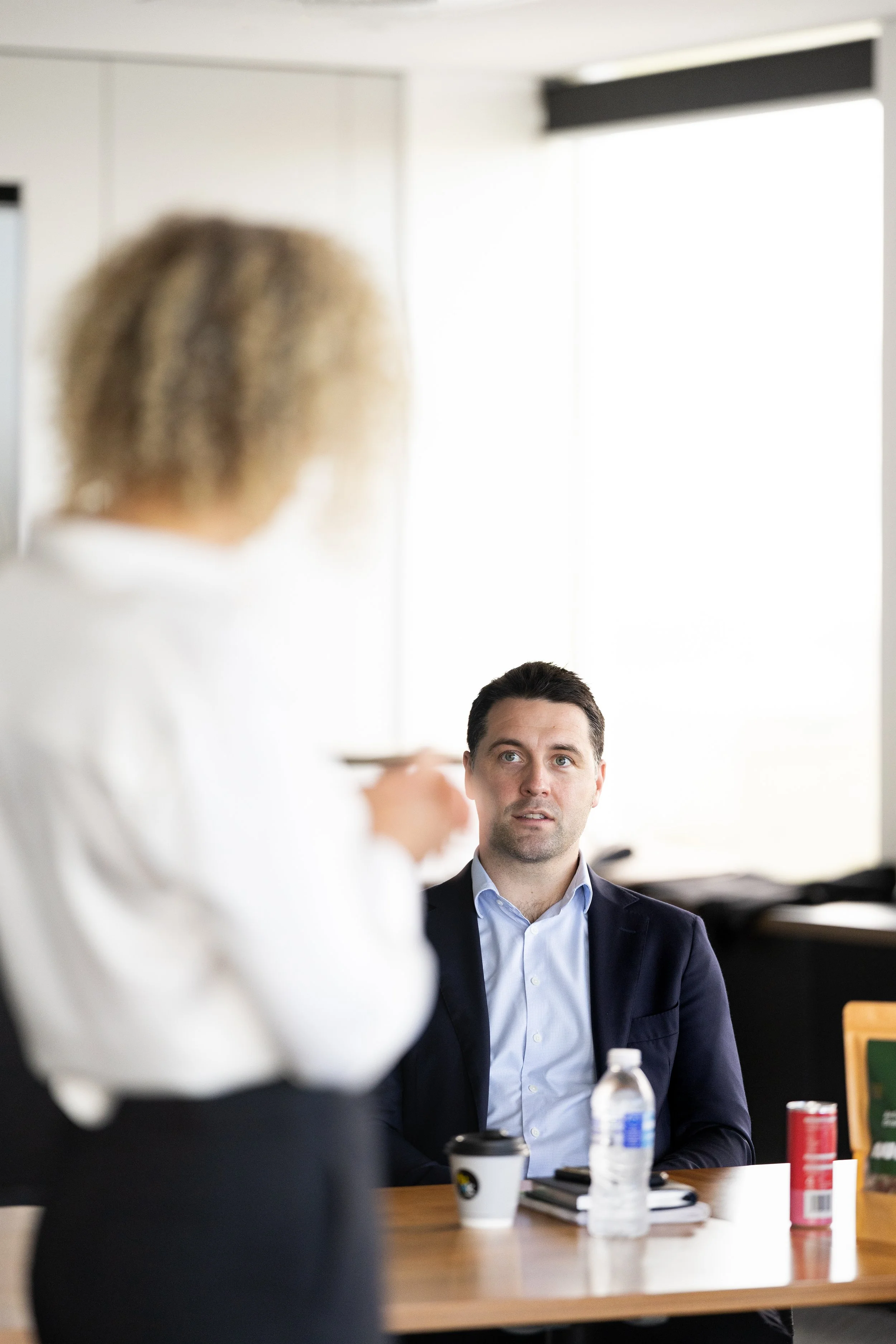 A business meeting with a man in a dark suit and light blue shirt listening to a woman with blonde hair presenting in the foreground.