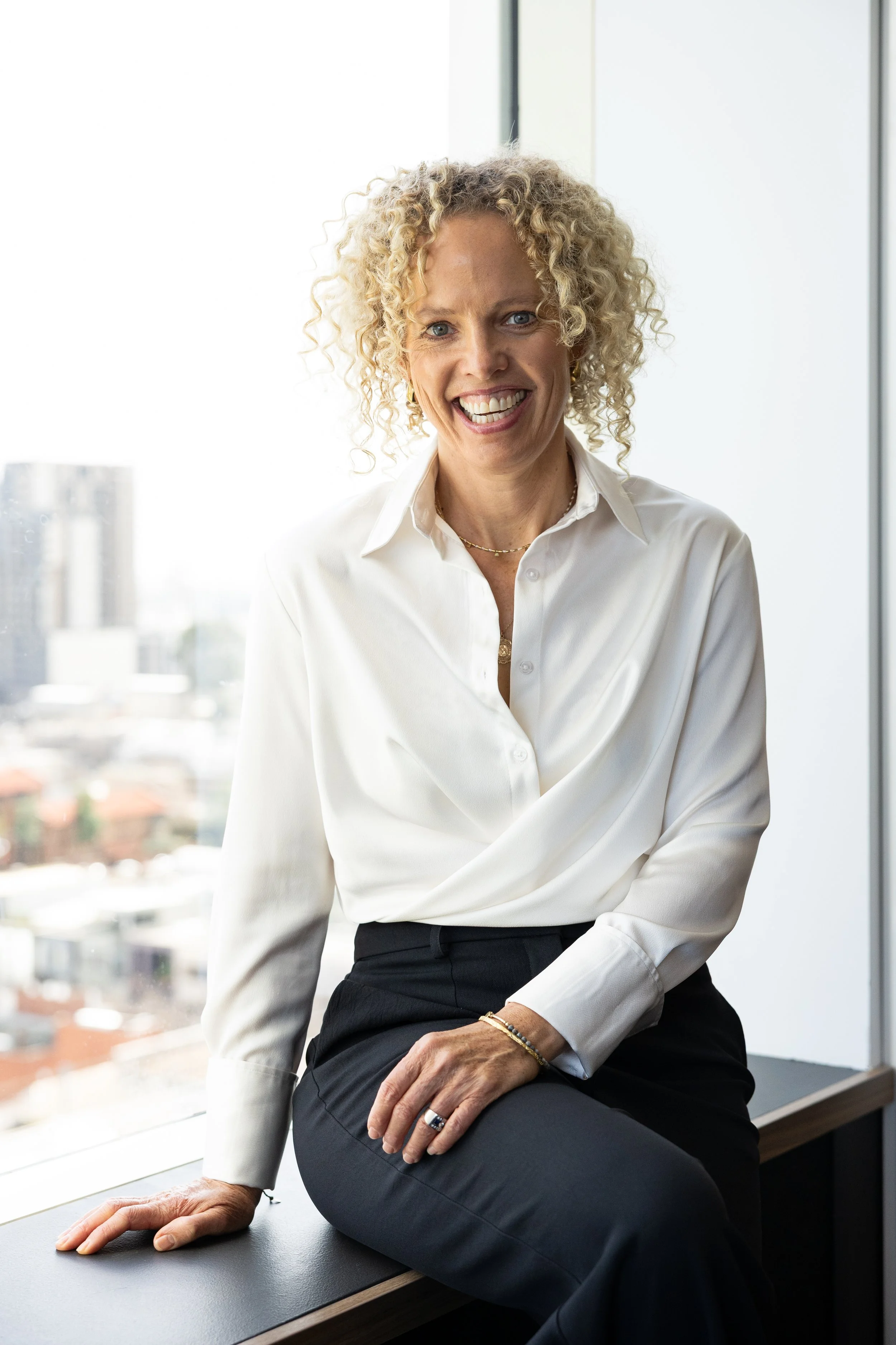 A woman with curly blonde hair smiling while sitting by a window in a business suit