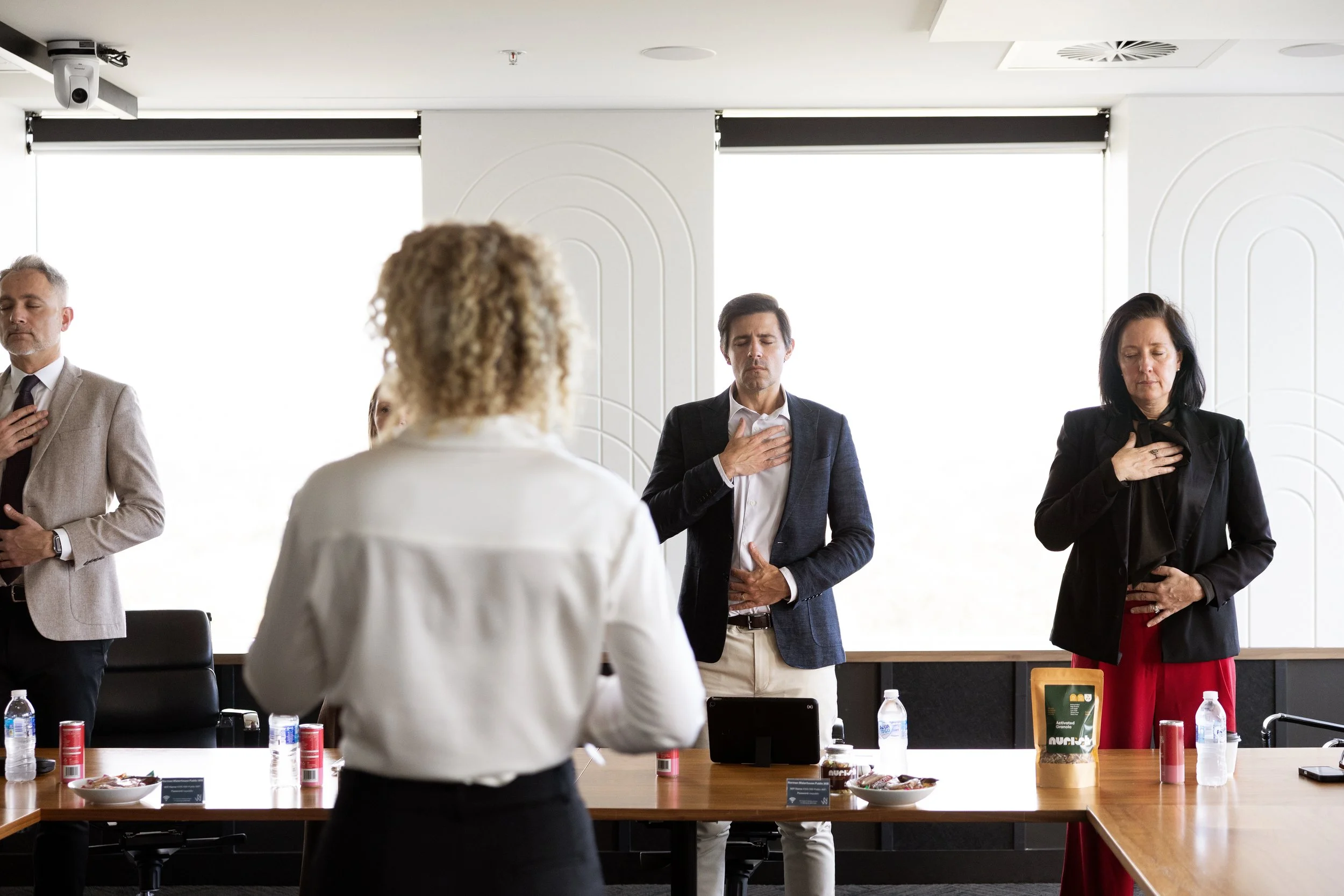 People standing with their right hands over their hearts in a meeting room during the national anthem.