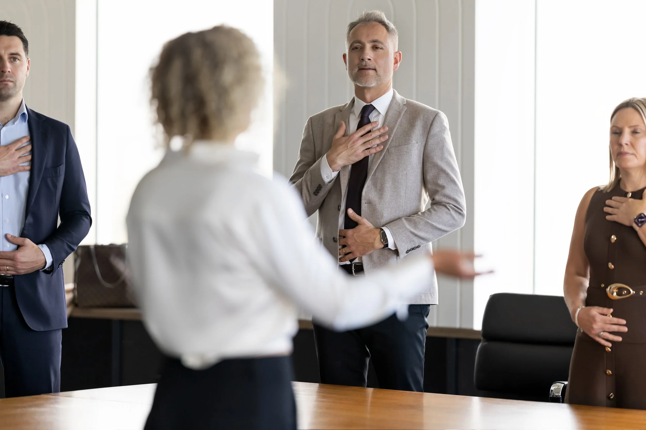 A group of professionals in a meeting with their right hands over their hearts, including a man in a light gray suit and a woman in a brown dress, standing in a bright room.