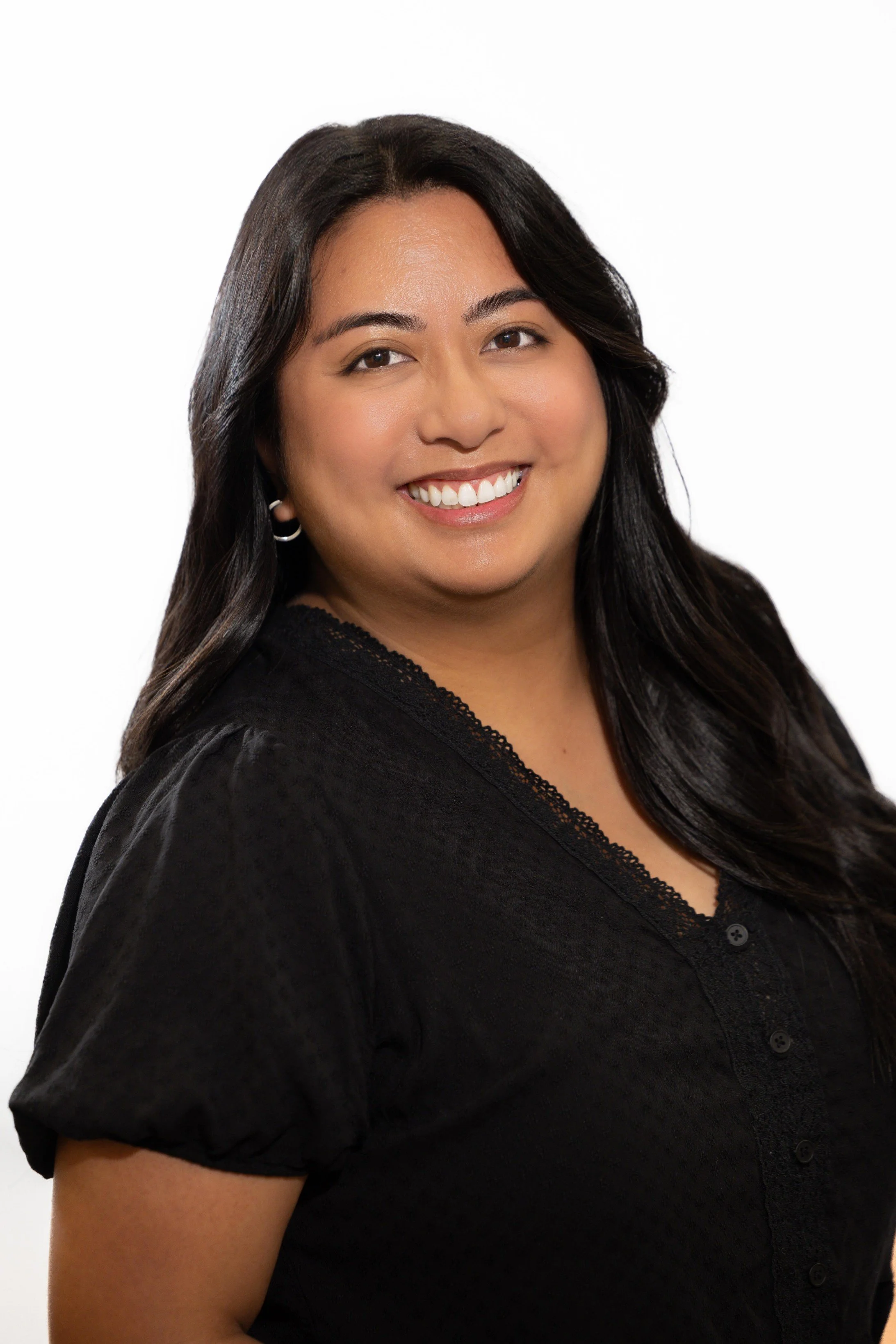 A woman with long dark hair and medium skin tone smiling, wearing a black top with lace trim and small hoop earrings, against a white background.