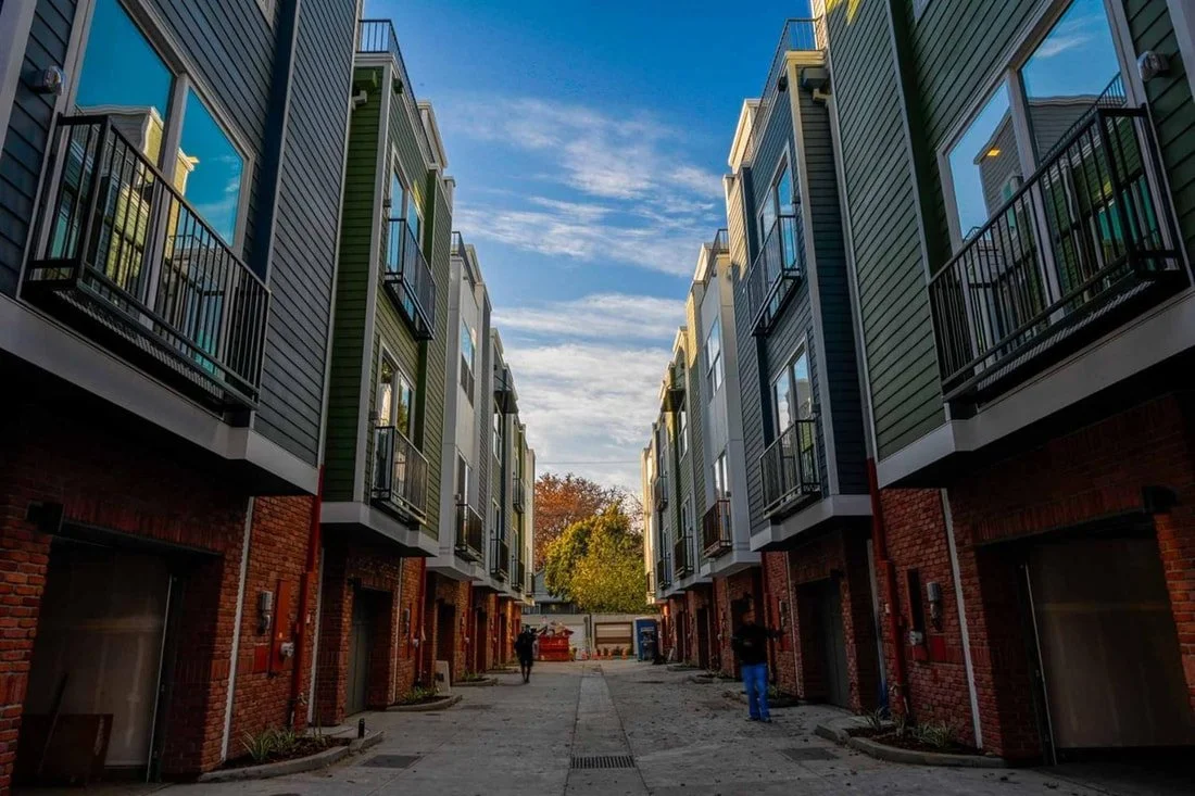 Two rows of modern multi-story residential buildings with brick ground floors and colorful upper floors, with balconies, lined along a paved alleyway with people walking.
