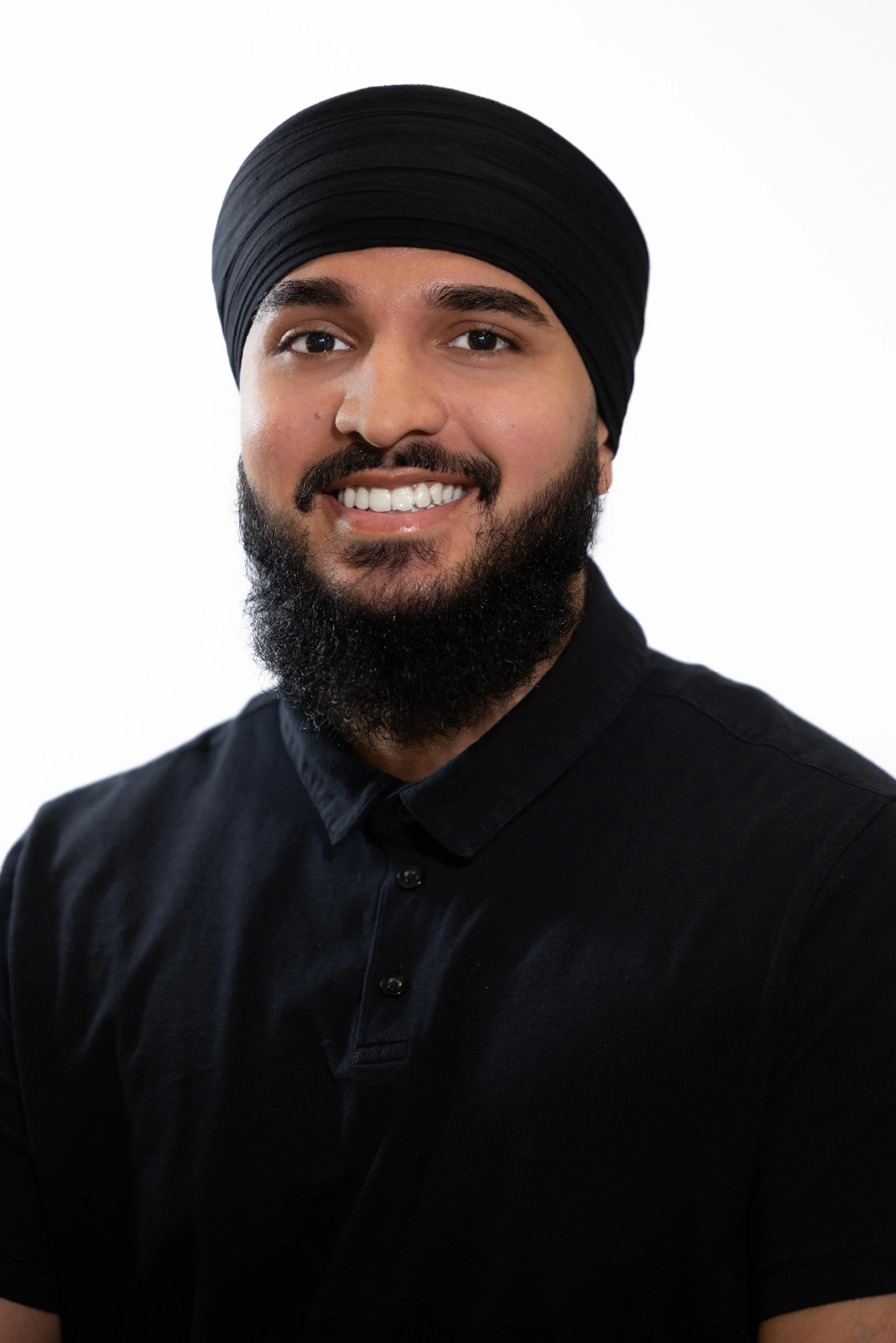 A smiling man with a dark beard, wearing a black turban and black shirt, against a white background.