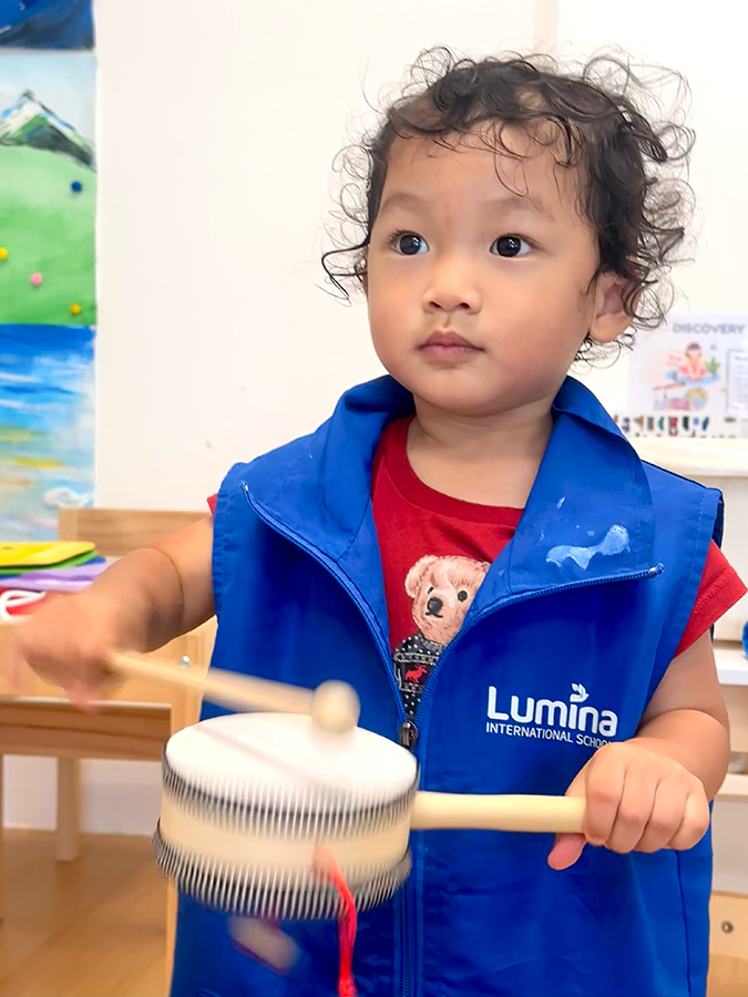 A young child with curly hair wearing a blue vest with Lumina International School written on it, holding a drumstick and playing a handheld drum in a classroom.