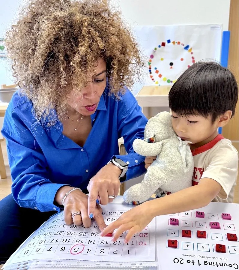 A man and a young girl sitting at a dining table, smiling as the girl writes or draws in a notebook with a pencil. There is a bowl of grapes on the table, and the man is standing behind her, leaning over to watch.