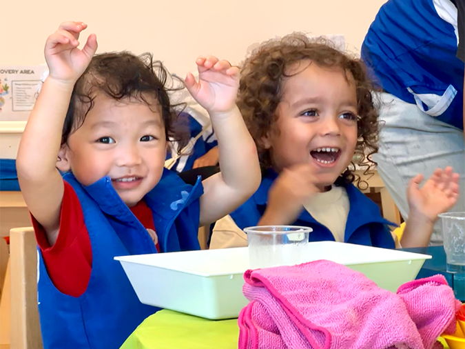 Two young children, one with curly hair and the other with straight hair, are smiling and raising their hands during an activity at a table, with pink cloth and clear containers in front of them in their classroom