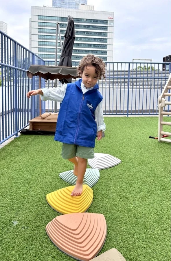 A young child balancing on colorful stepping stones on a rooftop play area with a cityscape background.