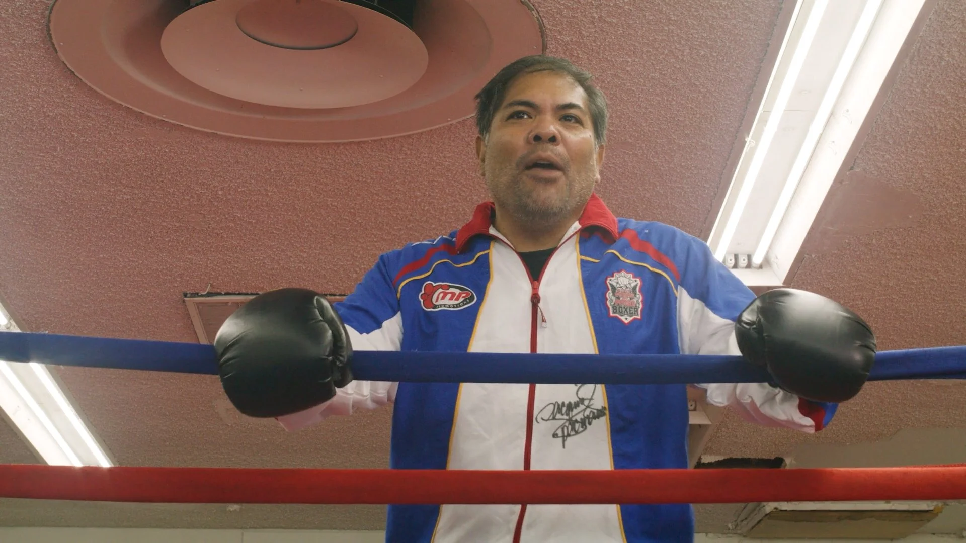 A man in boxing gloves leans on the ropes of a boxing ring, wearing a colorful jacket with logos and signature, in a gym with fluorescent lights and a pink ceiling.