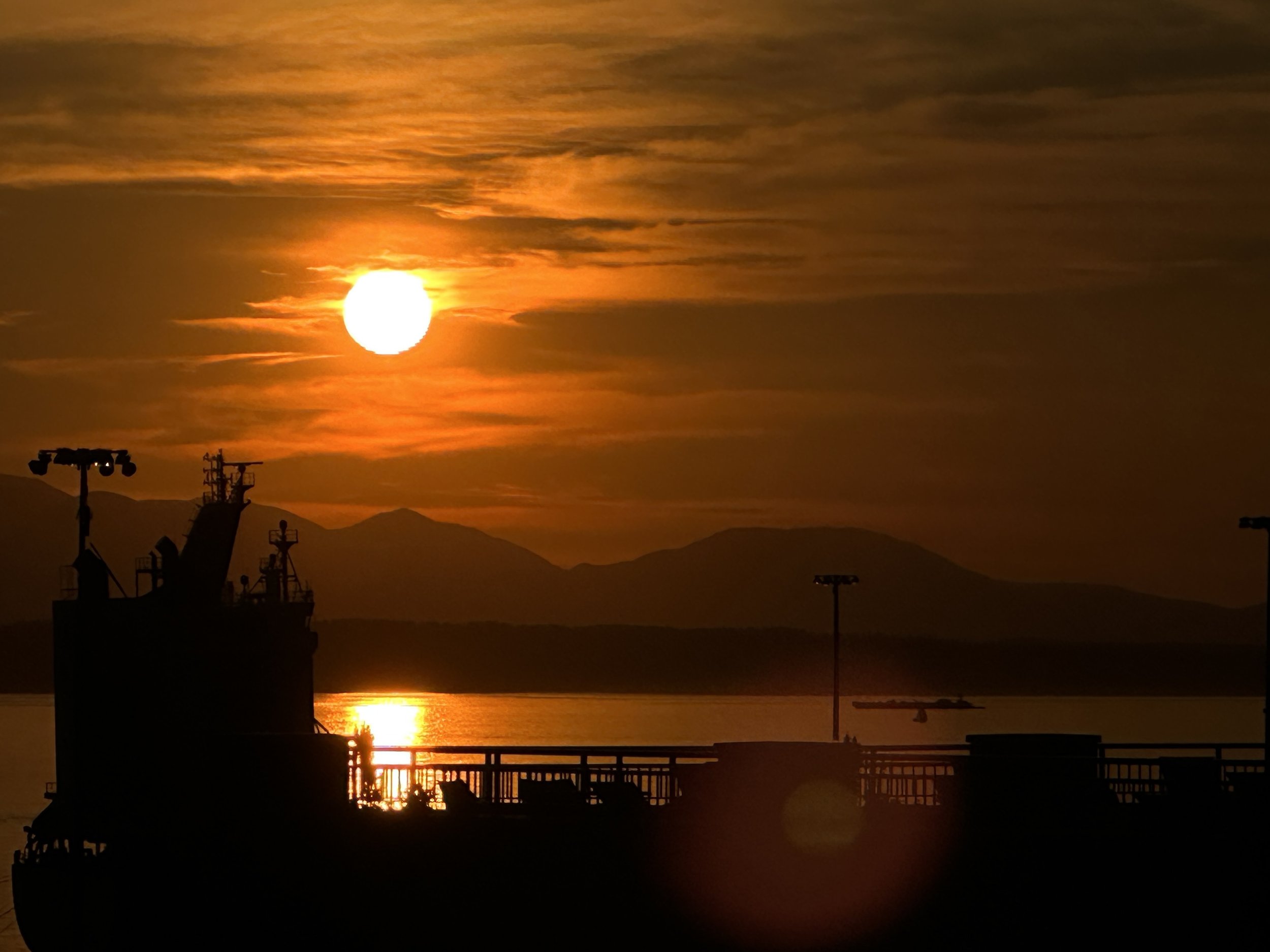 Sunset over a harbor with a ship silhouette and mountains in the distance.