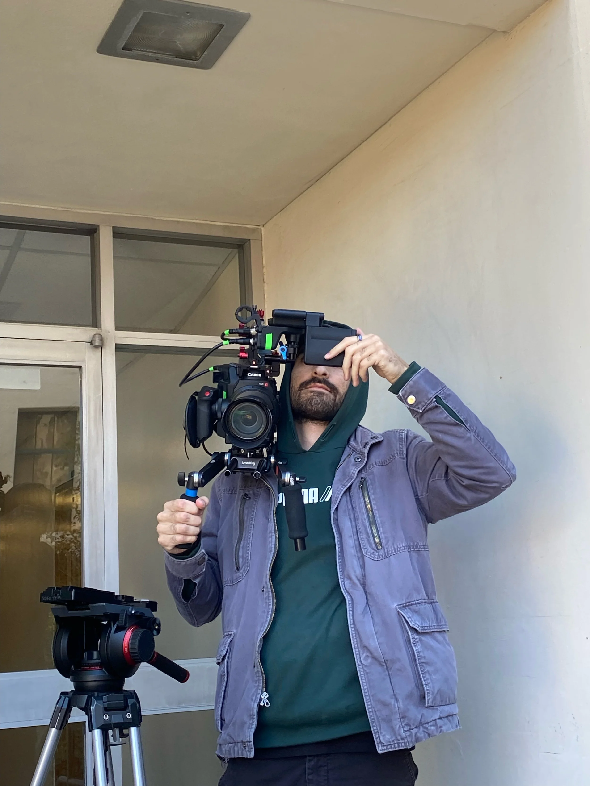 A man is holding a professional video camera on a tripod, wearing a jacket with a hoodie underneath, standing outside near a building with glass doors.