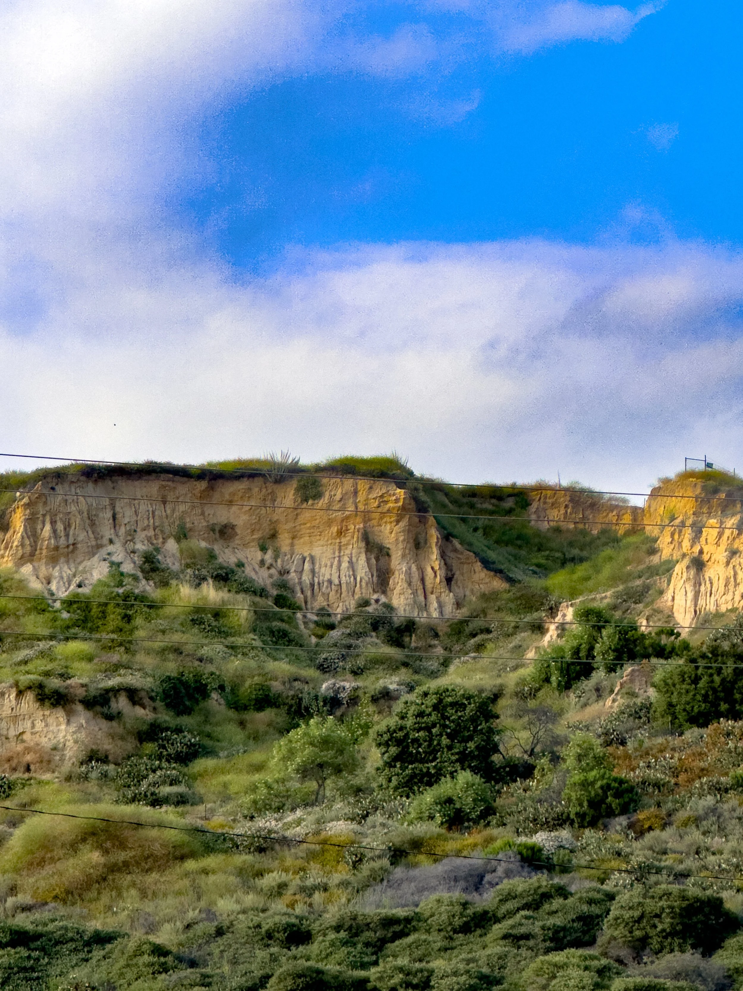 Hilly landscape with green vegetation, beige cliff formations, and a partly cloudy blue sky.
