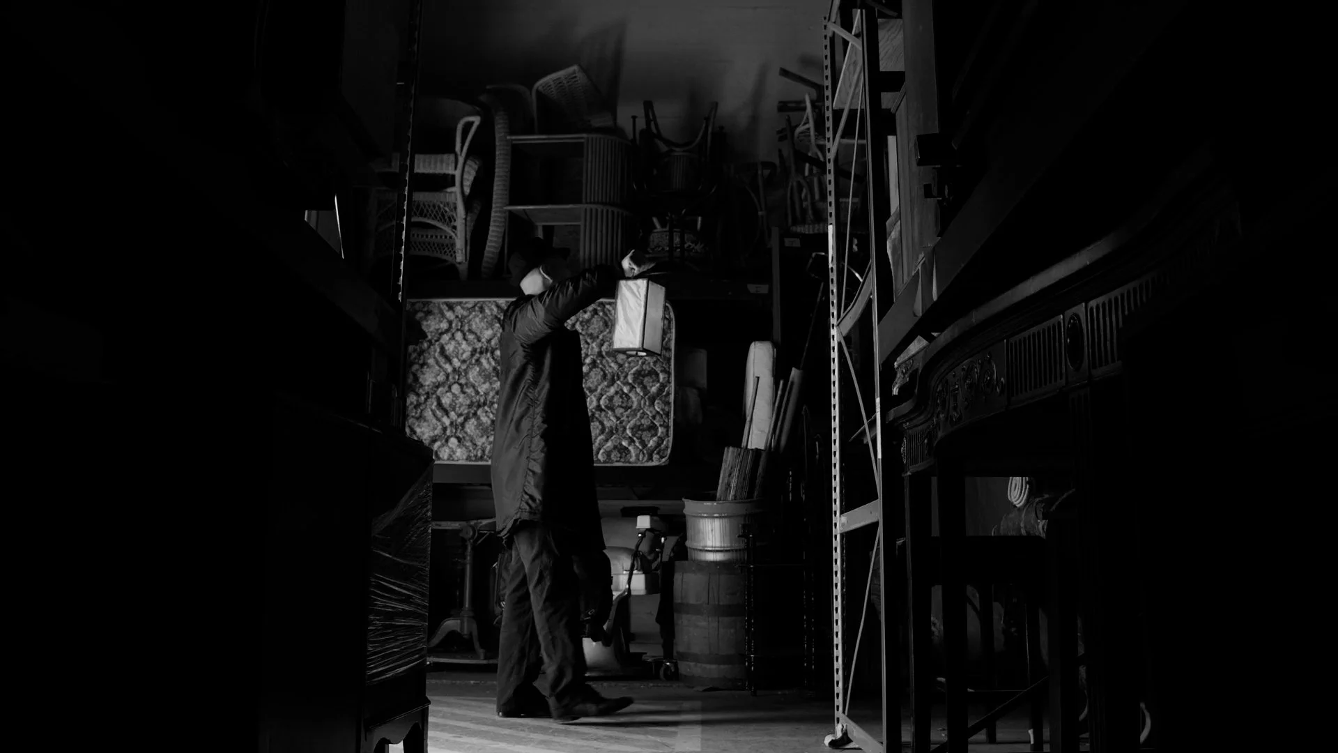 Person in a dark room reaching up to a high shelf with various furniture and boxes stacked around.