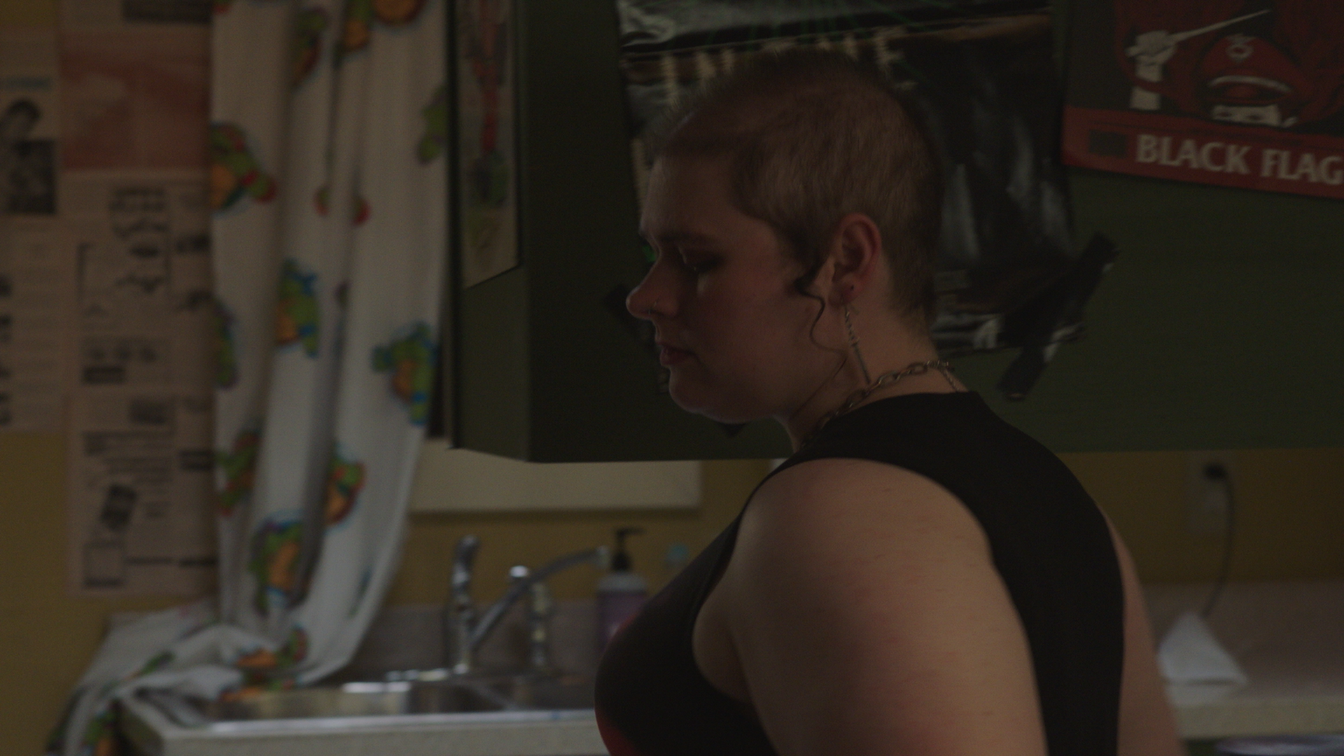 A woman with short hair and a tank top standing sideways in a kitchen, with a curtain featuring colorful cartoon characters, a sink, and posters on the wall in the background.