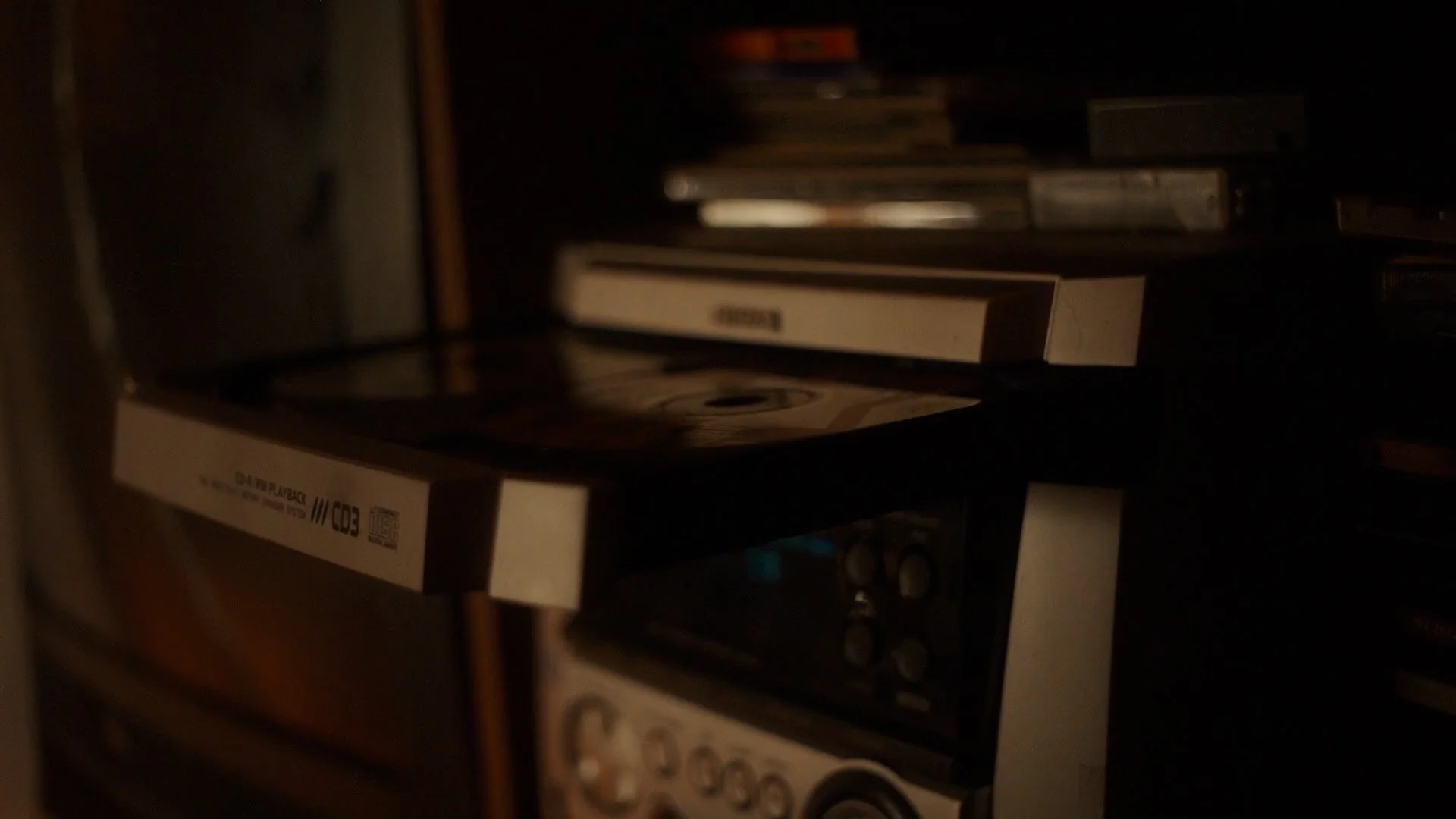 A dark, cluttered room with a CD player, a stack of CDs, and other electronic equipment.