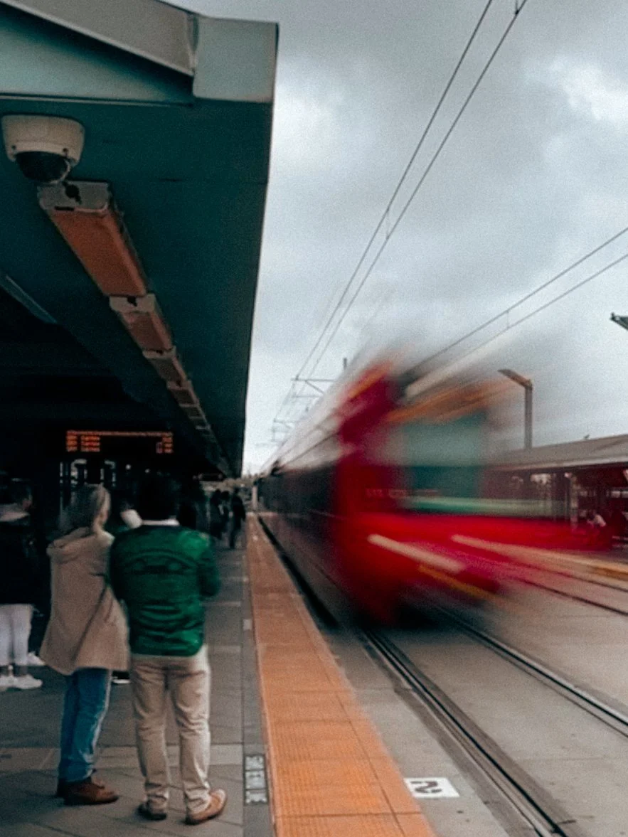 A train departing a station with a blurred motion effect and people waiting on the platform under an overcast sky.