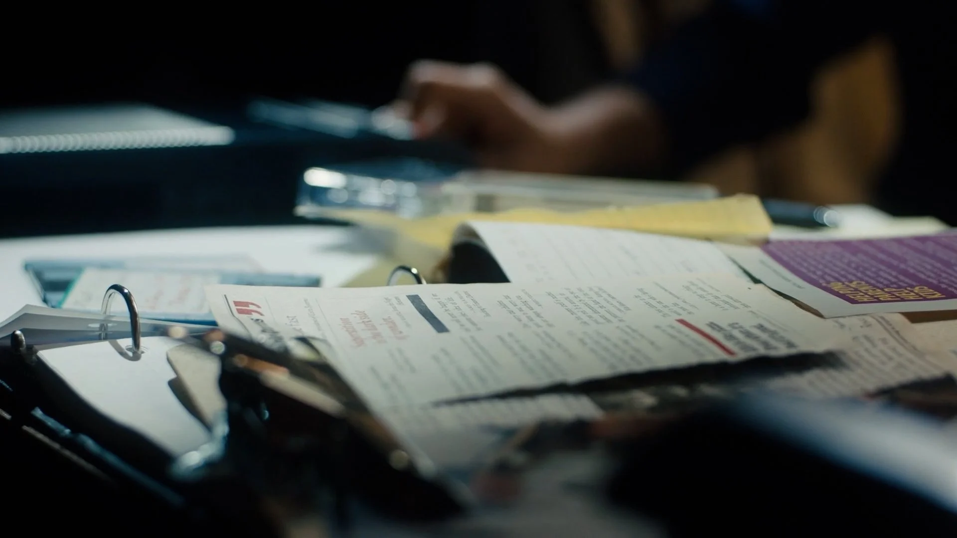 Desk cluttered with scattered papers, notebooks, and pens, with a person blurred in the background
