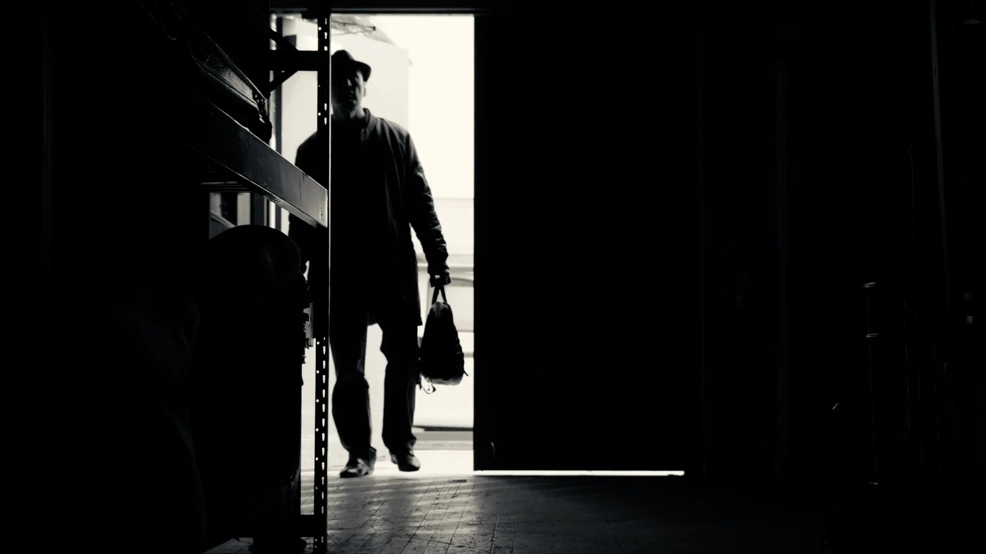 Silhouette of a man walking through a doorway carrying a bag in a dark room, with light coming from outside.