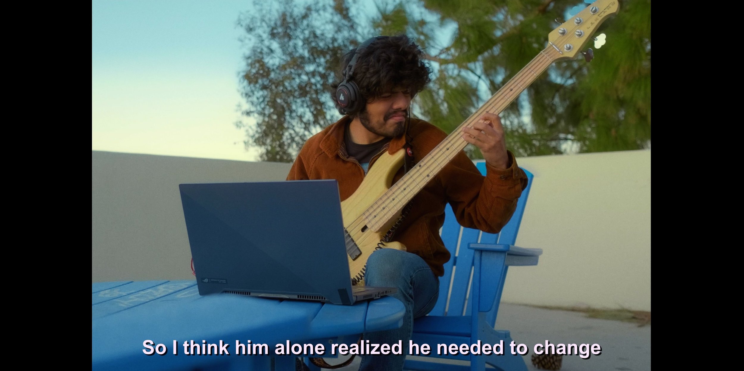 A young man wearing headphones playing a bass guitar while sitting at a blue table with a laptop, outdoors with trees in the background.