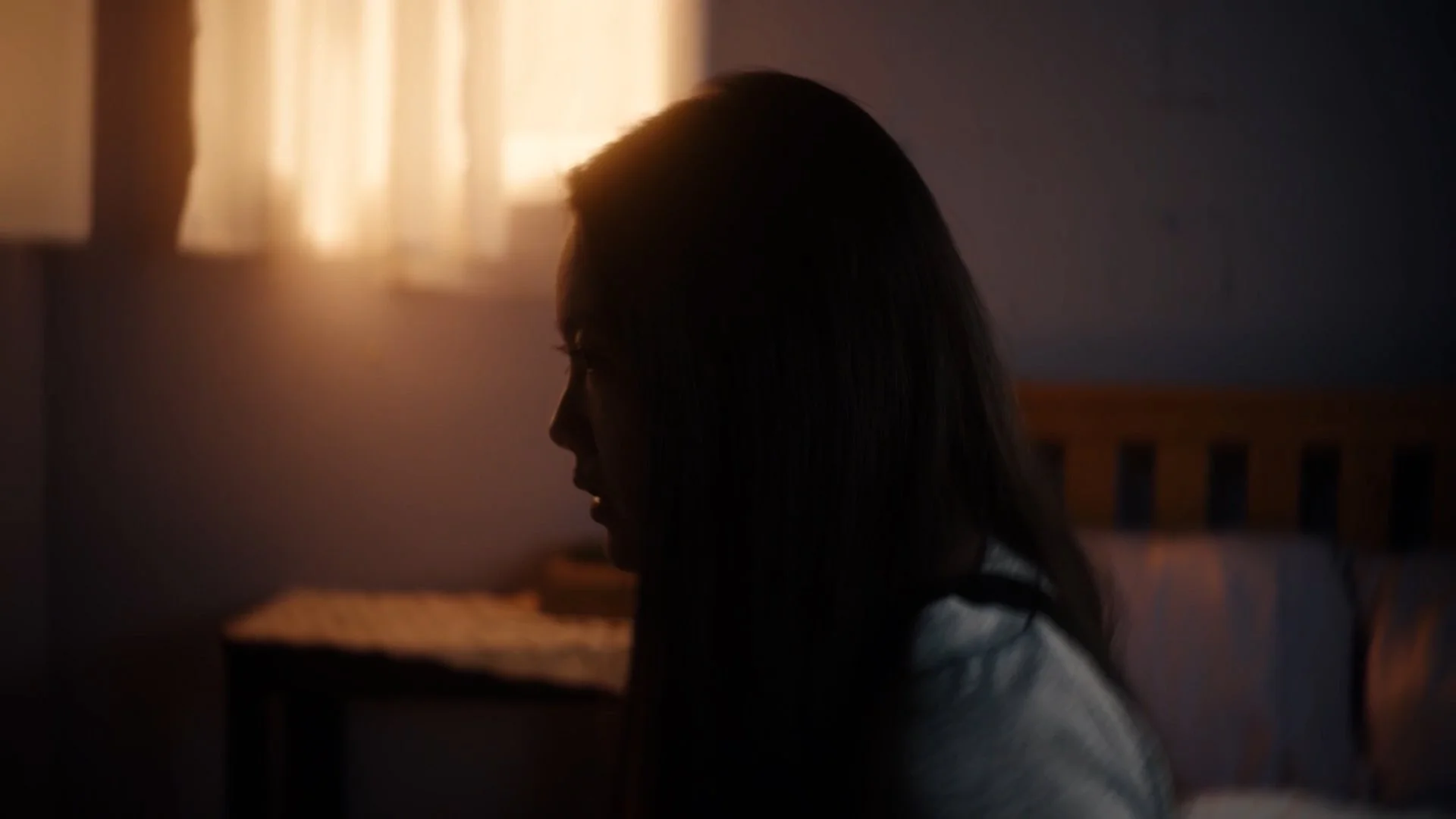 A woman sitting on a bed with her profile visible, illuminated by soft light coming through a window with curtains, in a dimly lit room.